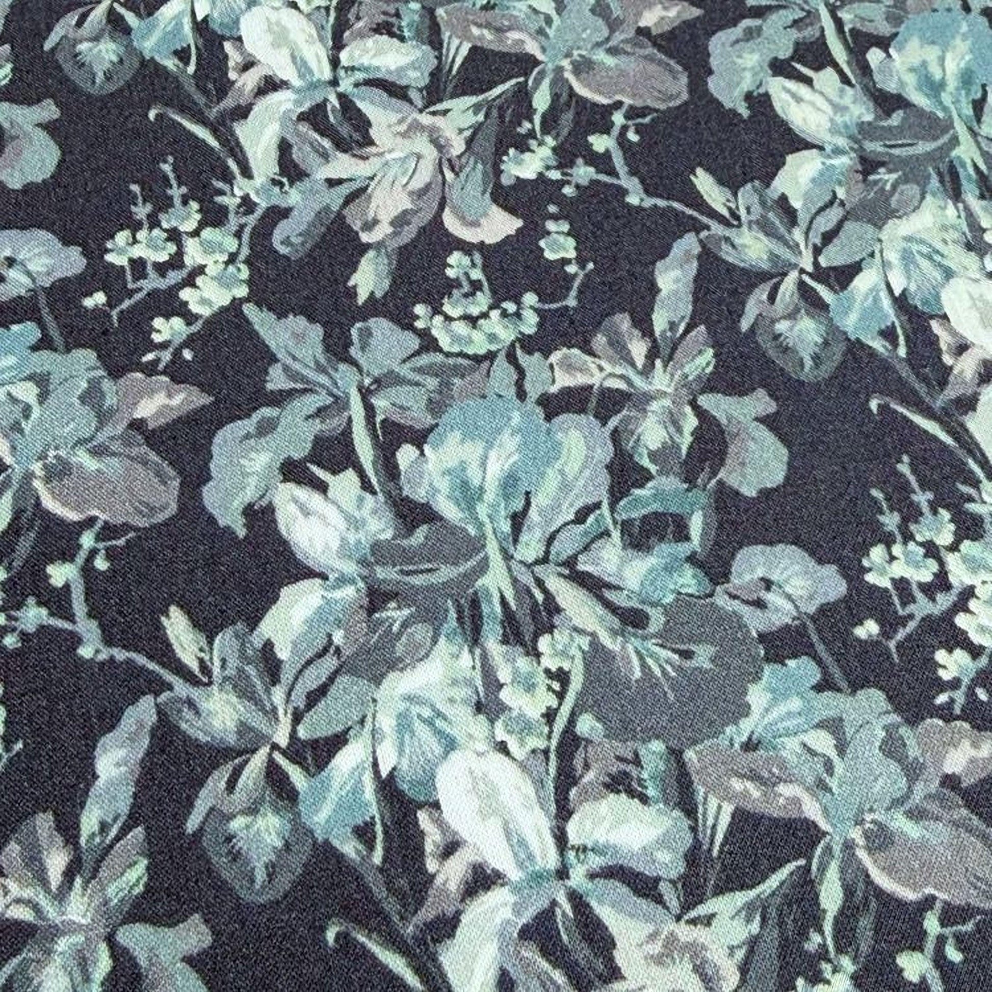 a close-up view of a fabric with a floral pattern in shades of blue and gray. The pattern consists of various flowers and leaves, creating a visually appealing and intricate design. The fabric appears to be dark in color, which contrasts with the lighter shades of the flowers and leaves. The image does not contain any text or other objects, and the focus is solely on the floral pattern on the fabric.