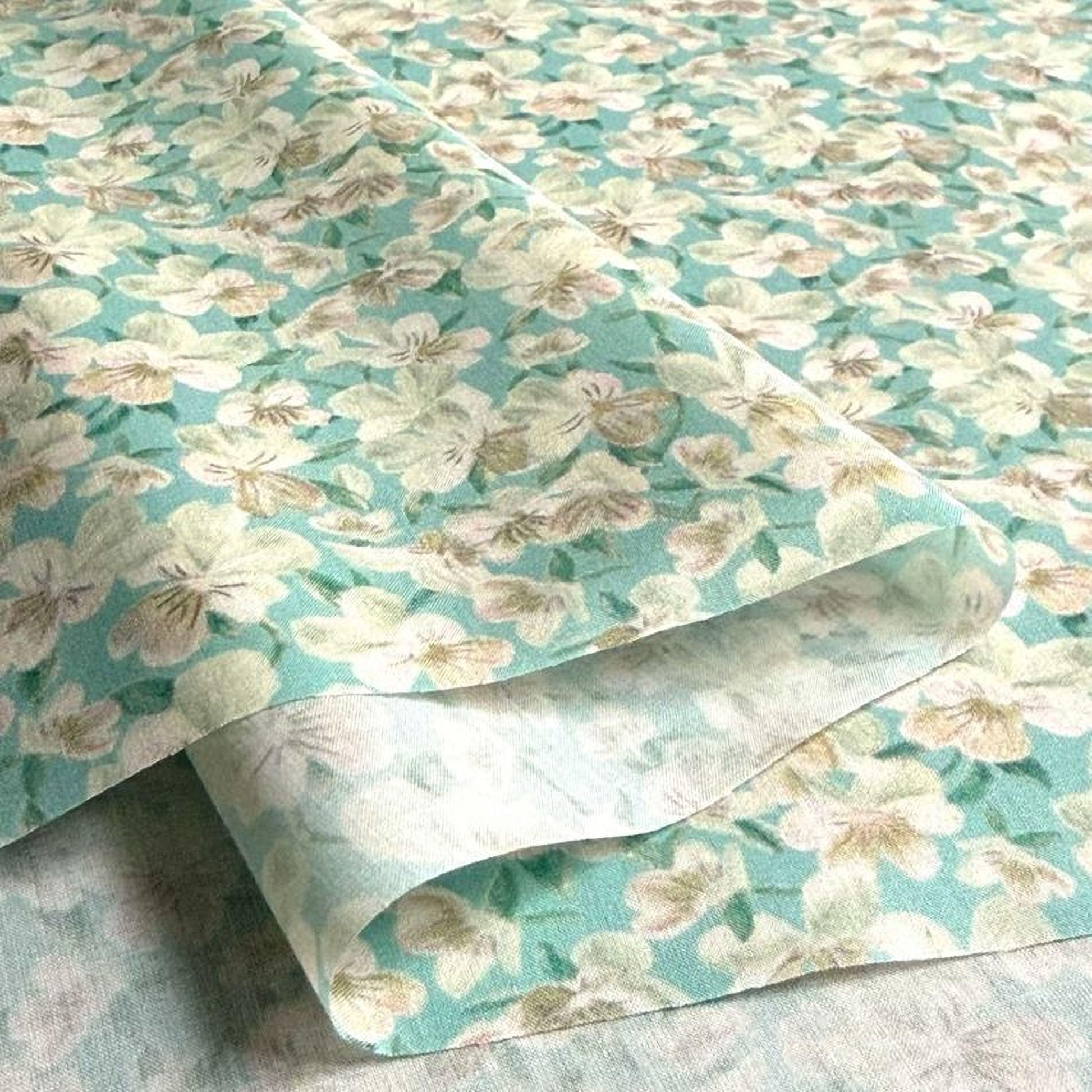 a close-up view of a fabric with a floral pattern in shades of green and white. The fabric appears to be folded or rolled, revealing the intricate design of the flowers.