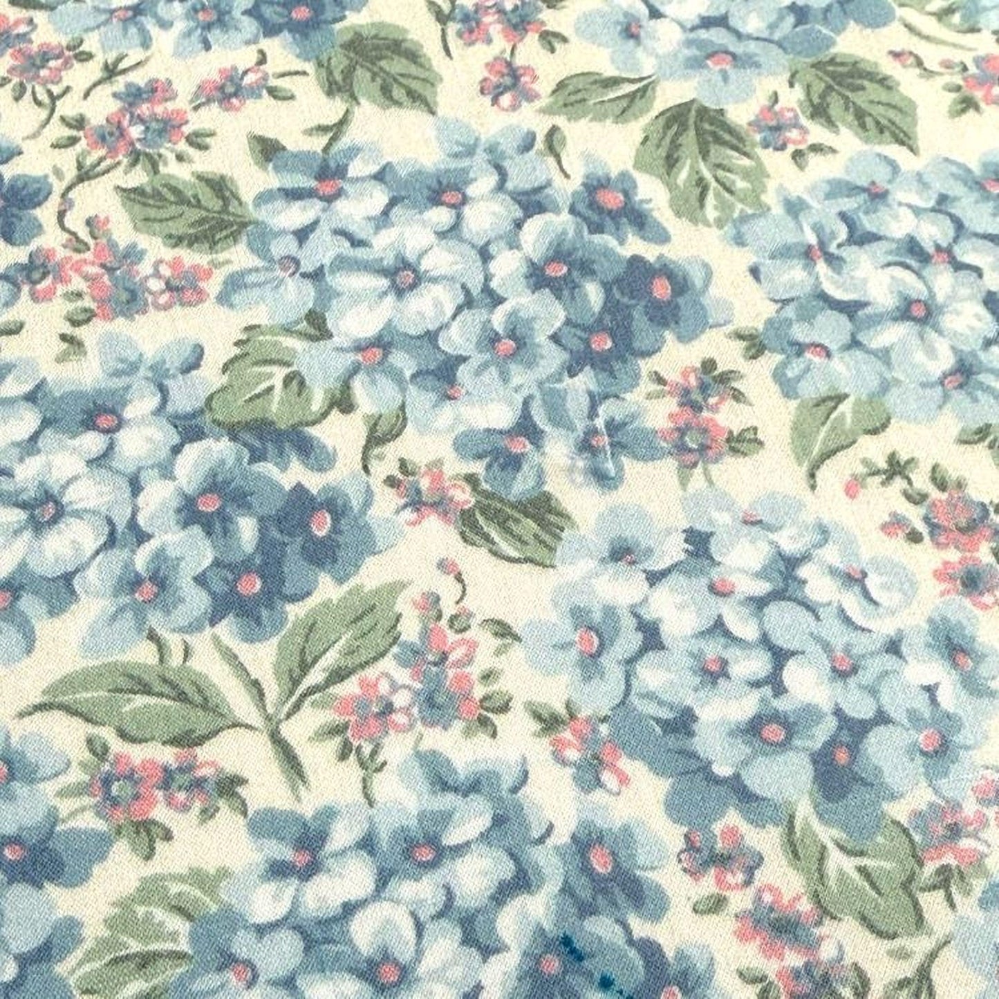 a close-up view of a fabric with a floral pattern. The fabric is predominantly blue and white, with scattered pink and green accents. The pattern consists of various blue flowers and leaves, interspersed with pink and green accents. The fabric appears to be a light-colored material, possibly cotton or linen, with a textured surface.
