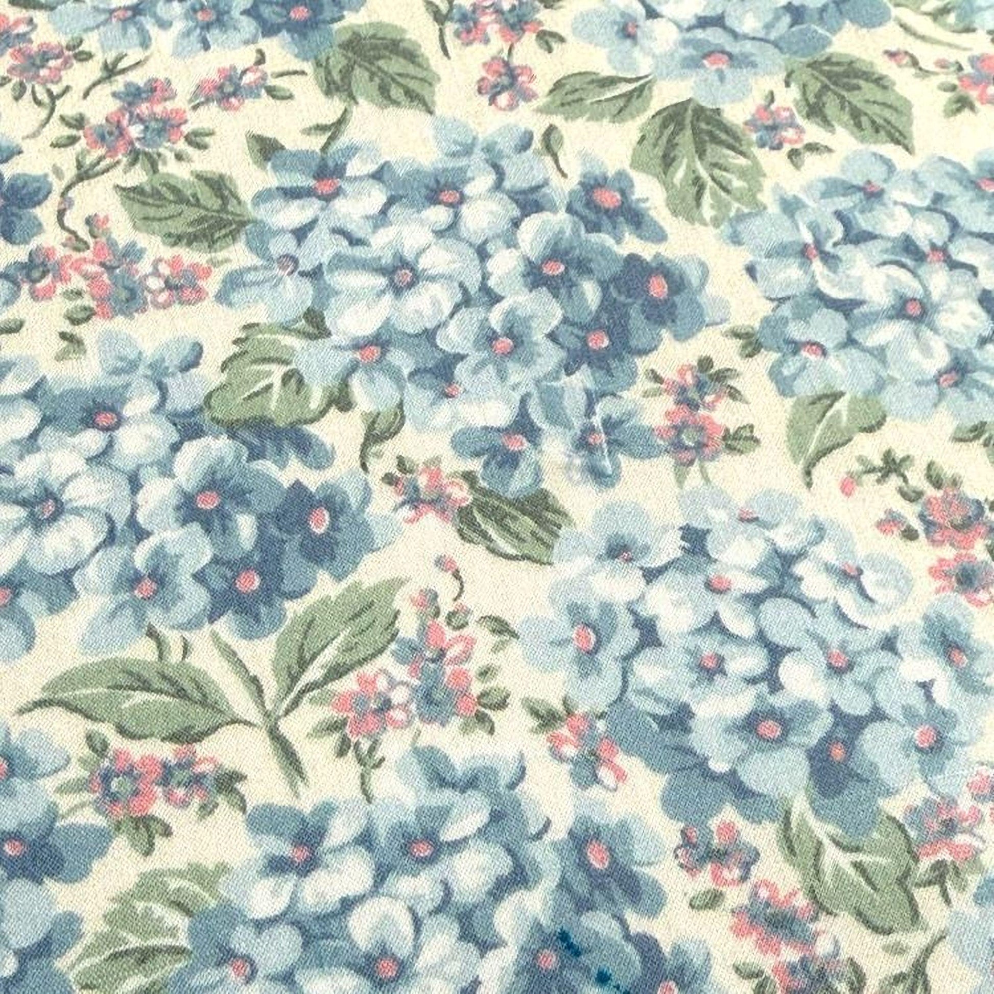 a close-up view of a fabric with a floral pattern. The fabric is predominantly blue and white, with scattered pink and green accents. The pattern consists of various blue flowers and leaves, interspersed with pink and green accents. The fabric appears to be a light-colored material, possibly cotton or linen, with a textured surface.