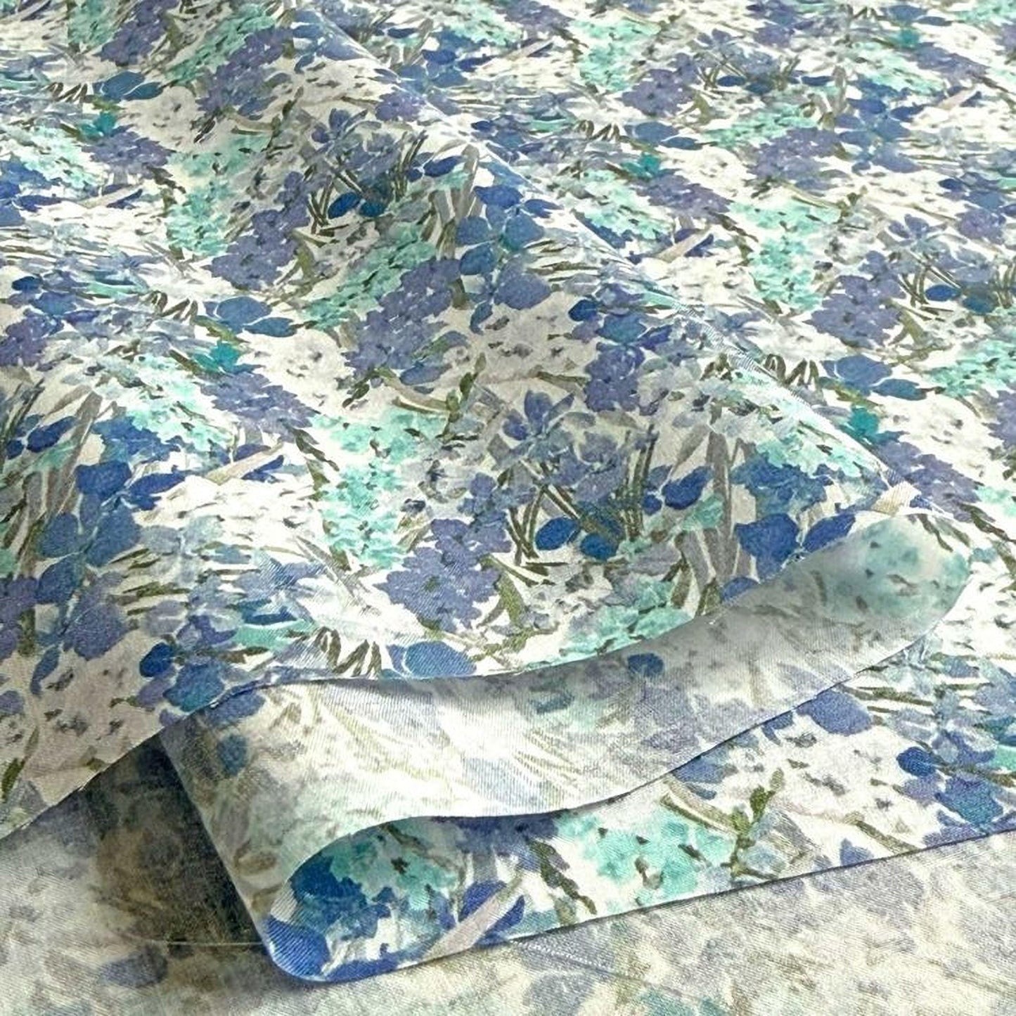 a close-up view of a fabric with a floral pattern in shades of blue and green. The fabric appears to be folded or rolled, revealing the intricate design of the flowers and leaves.