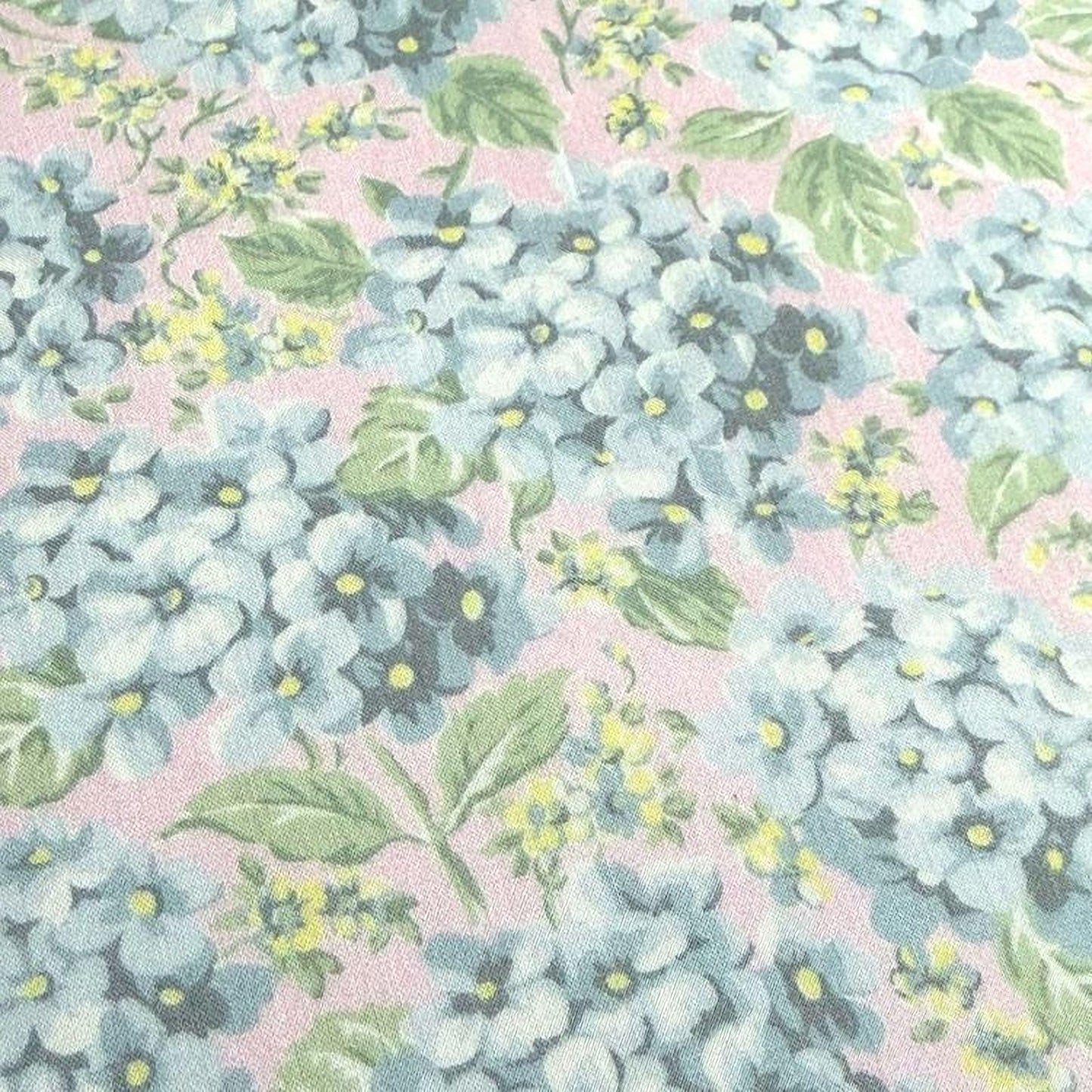 a close-up view of a fabric with a floral pattern. The fabric has a mix of blue and green flowers, along with green leaves, creating a vibrant and colorful design. The flowers appear to be in various stages of bloom, adding a dynamic element to the overall pattern. The fabric's texture and color combination suggest it could be used for clothing or home decor items.