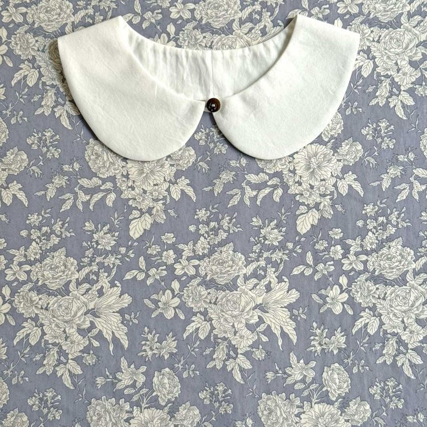 A white collar with a button is displayed against a background of a floral patterned fabric.