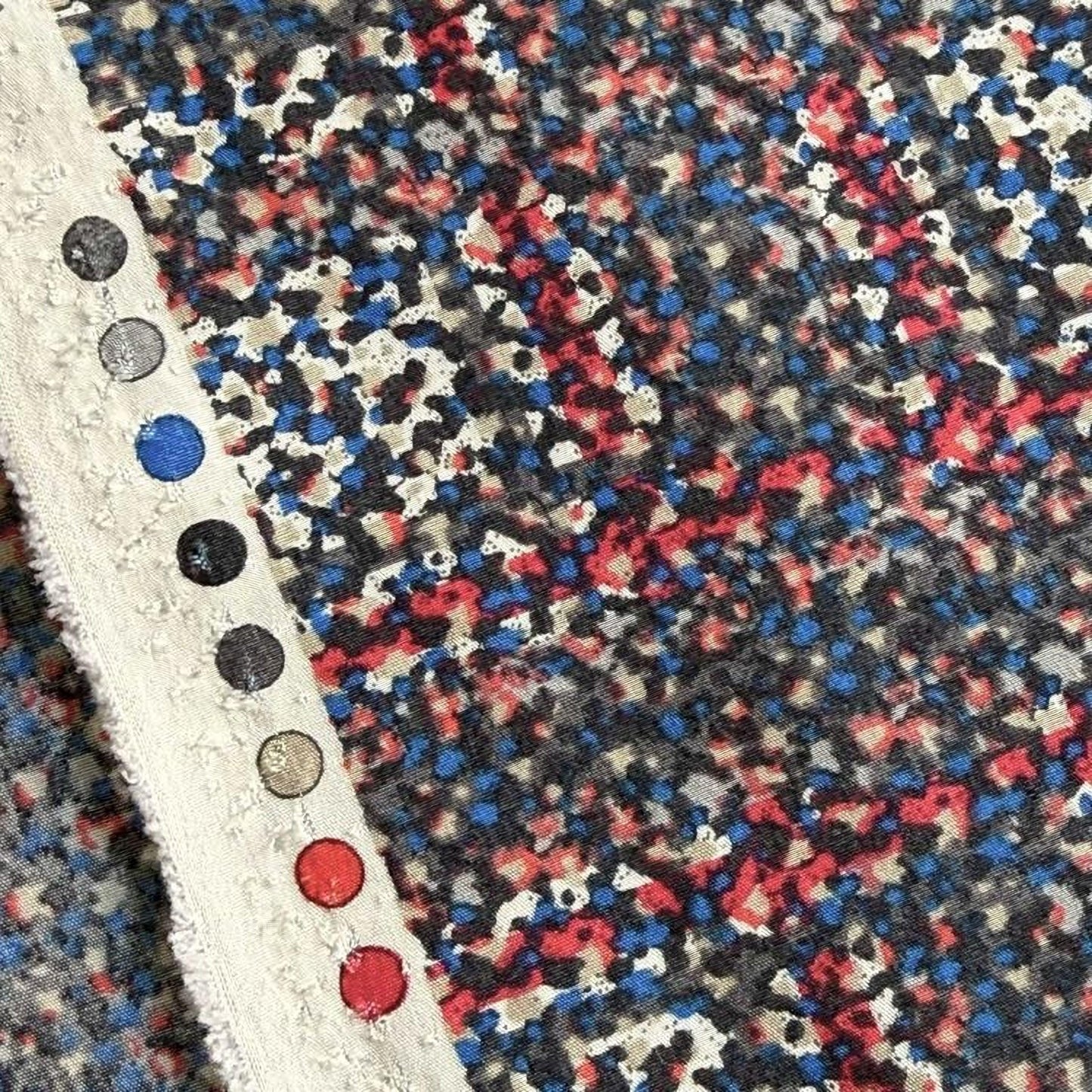 a close-up view of a fabric with a multicolored, abstract pattern. The fabric appears to be a piece of clothing or textile, with a white trim or border. The pattern consists of various shades of red, blue, and black, creating a vibrant and dynamic visual effect.