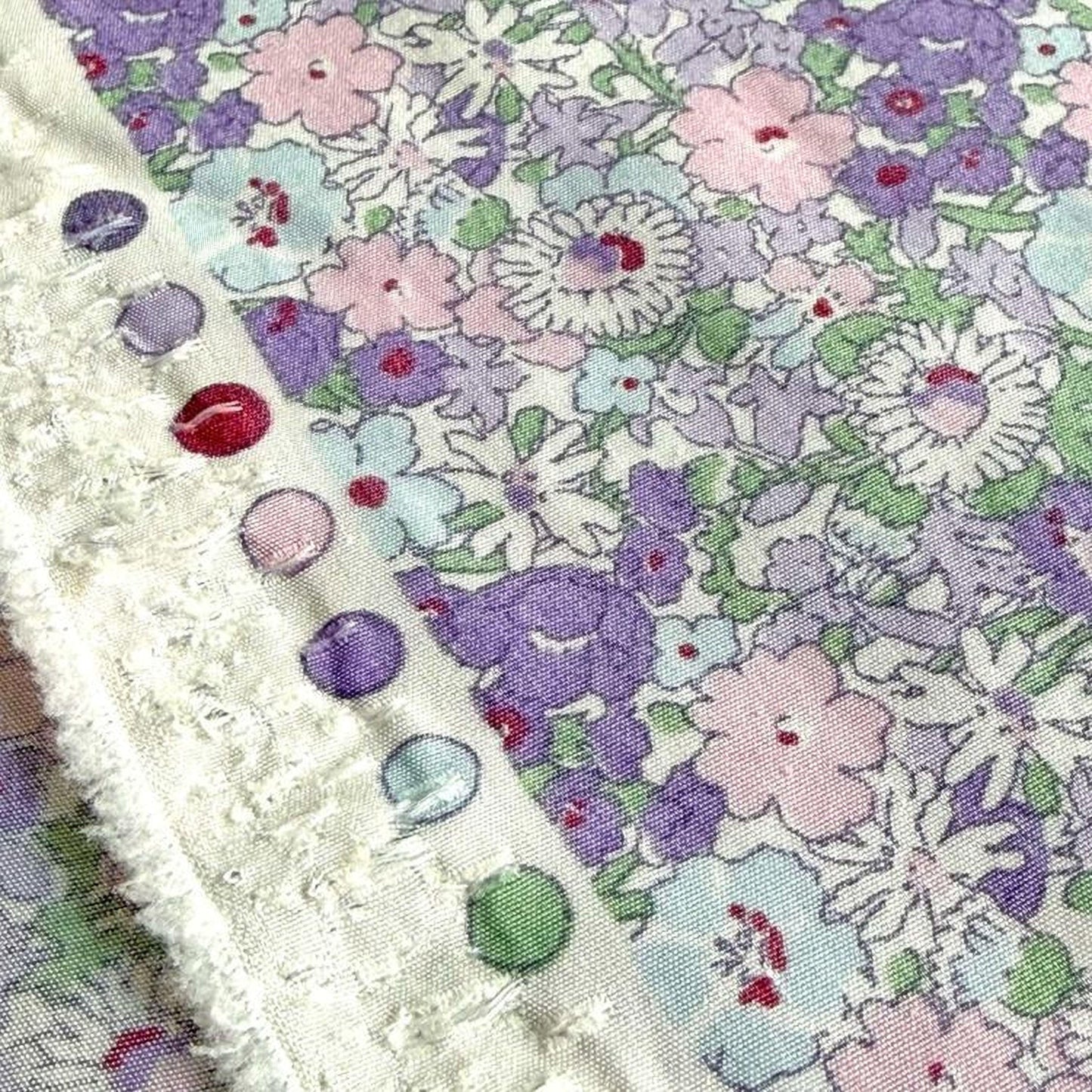 a close-up view of a fabric with a floral pattern, featuring a variety of colors including purple, pink, and white. The fabric appears to be a piece of clothing or textile, with a fringed edge and a row of colorful buttons visible on the edge.