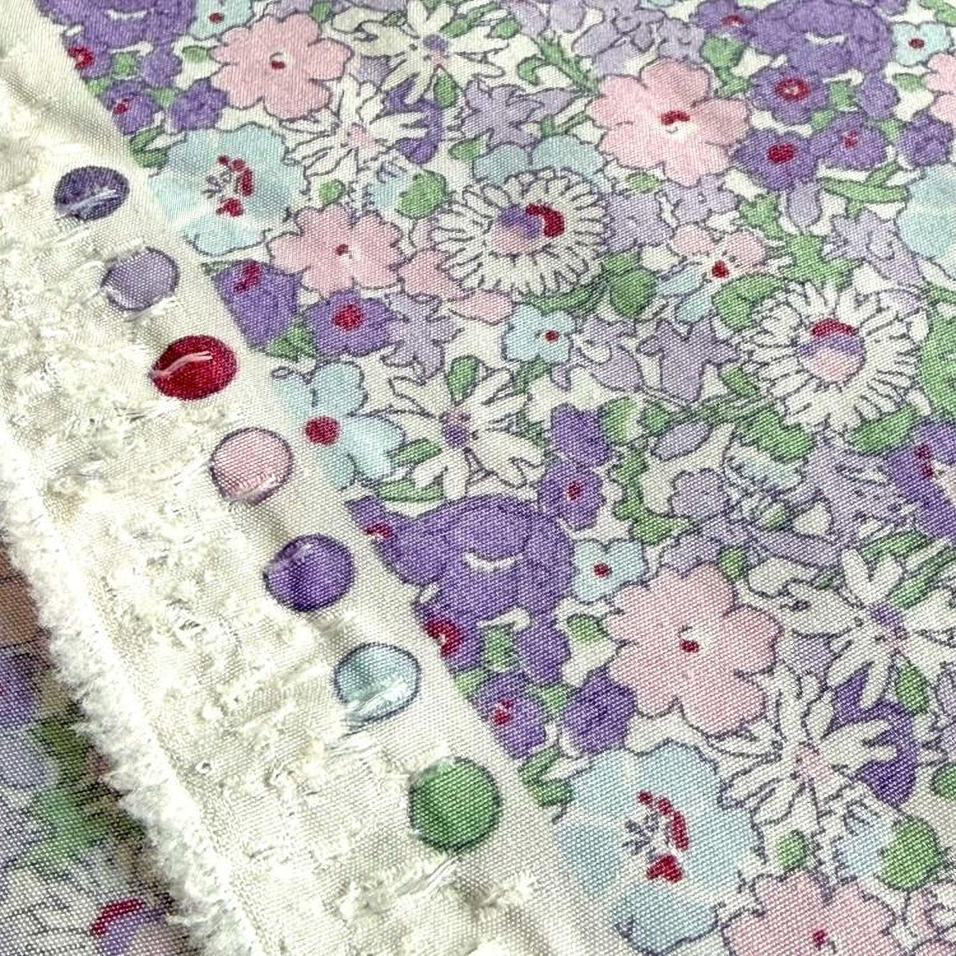 a close-up view of a fabric with a floral pattern, featuring a variety of colors including purple, pink, and white. The fabric appears to be a piece of clothing or textile, with a fringed edge and a row of colorful buttons visible on the edge.