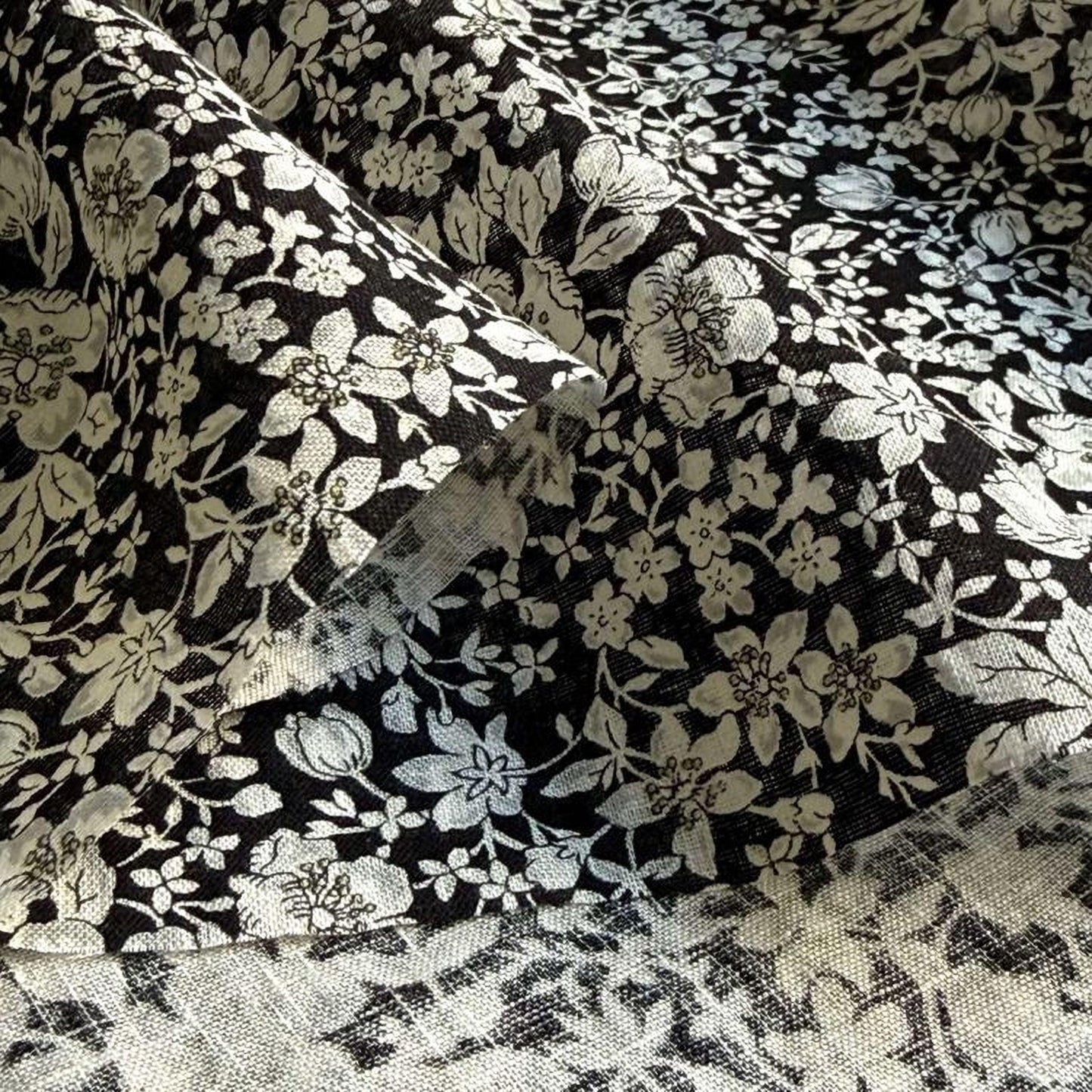 a close-up view of a fabric with a black and white floral pattern. The fabric appears to be made of a soft, textured material, possibly cotton or linen. The pattern consists of various white flowers and leaves, interspersed with black silhouettes, creating a visually striking contrast. The fabric is folded and crumpled, giving it a slightly wrinkled appearance.