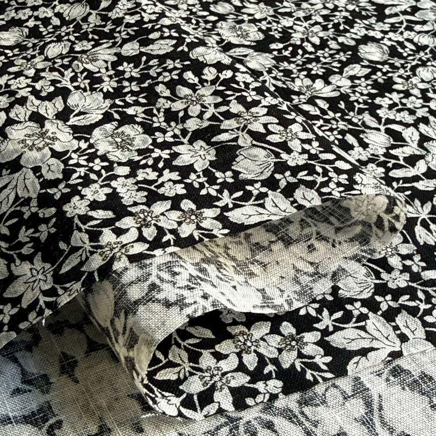 a close-up view of a fabric with a black and white floral pattern. The fabric appears to be made of a soft, textured material, possibly cotton or linen. The pattern consists of various flowers and leaves in different sizes and shapes, creating a visually appealing and intricate design. The image does not contain any text or other objects, and the focus is solely on the fabric and its floral pattern.