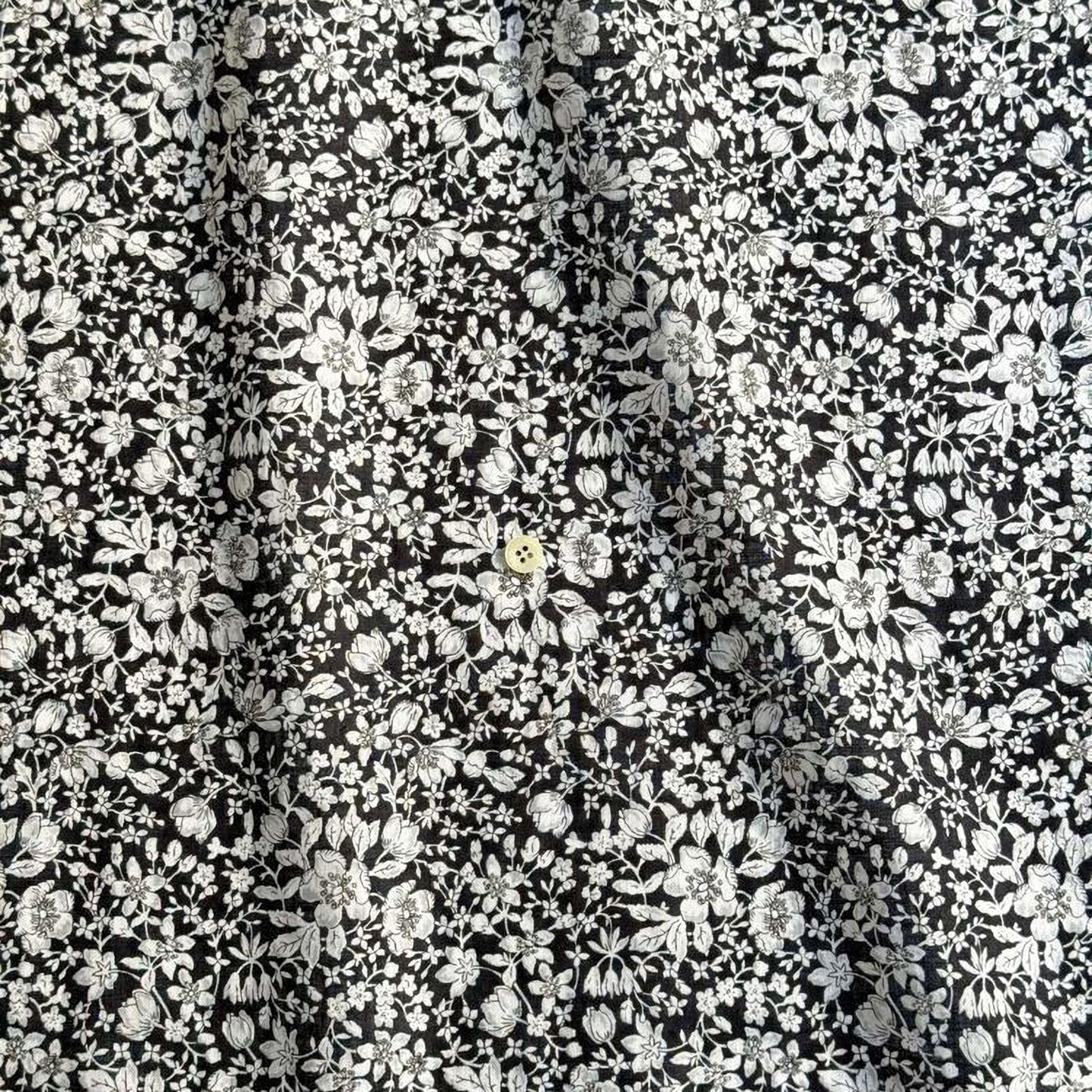 The image is a close-up of a fabric with a black and white floral pattern. The fabric appears to be made of a material with a floral design, possibly cotton or linen. The pattern consists of numerous small white flowers and leaves arranged in a repeating design across the fabric's surface. The image does not contain any text or other discernible objects or actions.