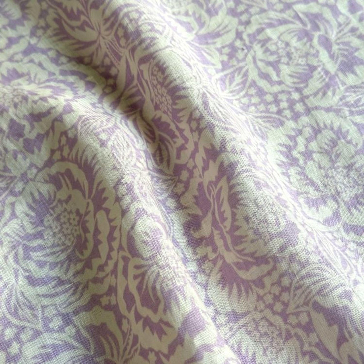 a close-up view of a fabric with a repeating pattern of purple and white floral designs. The fabric appears to be made of a soft, textured material, and the pattern is intricate and detailed.