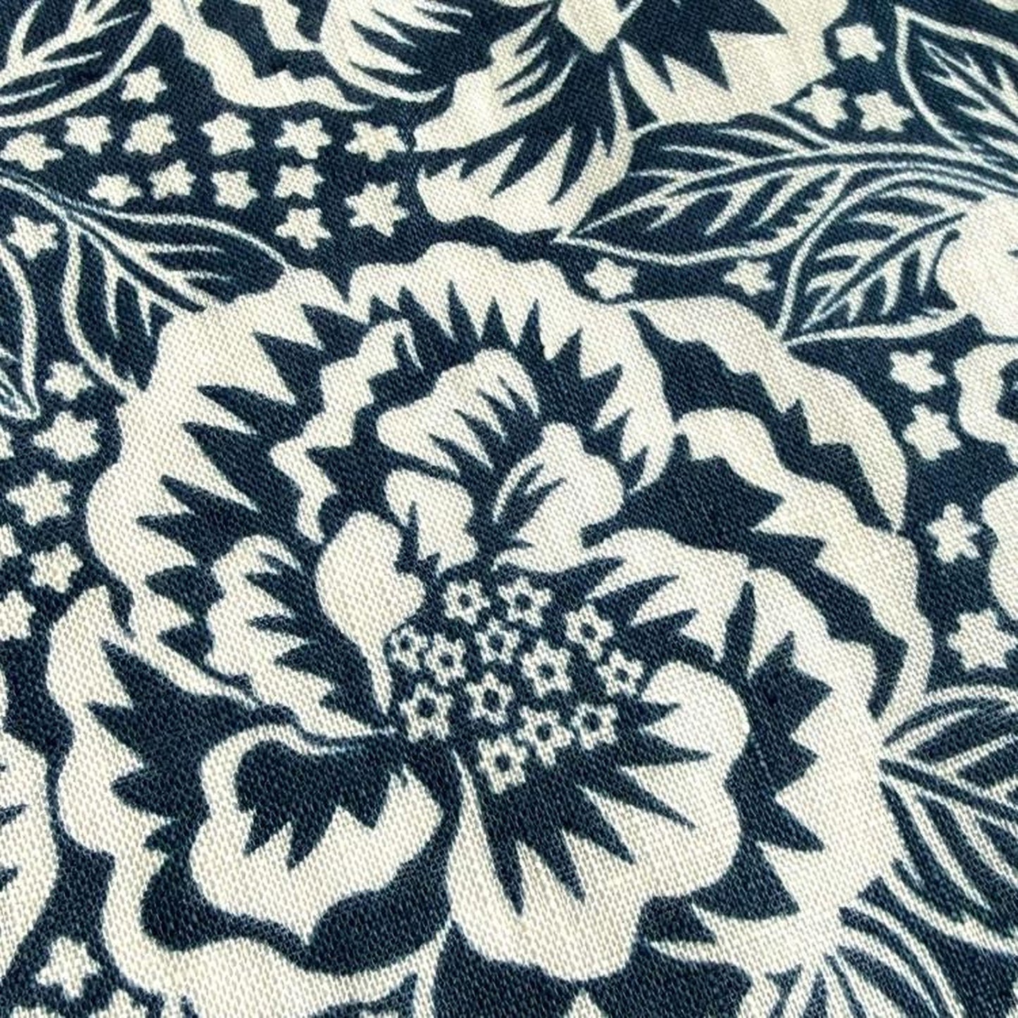 a close-up view of a fabric with a black and white floral pattern. The pattern consists of large, stylized flowers with intricate details and small white flowers scattered throughout the design. The fabric appears to be a dark-colored material, possibly a type of cotton or linen, with a textured surface.