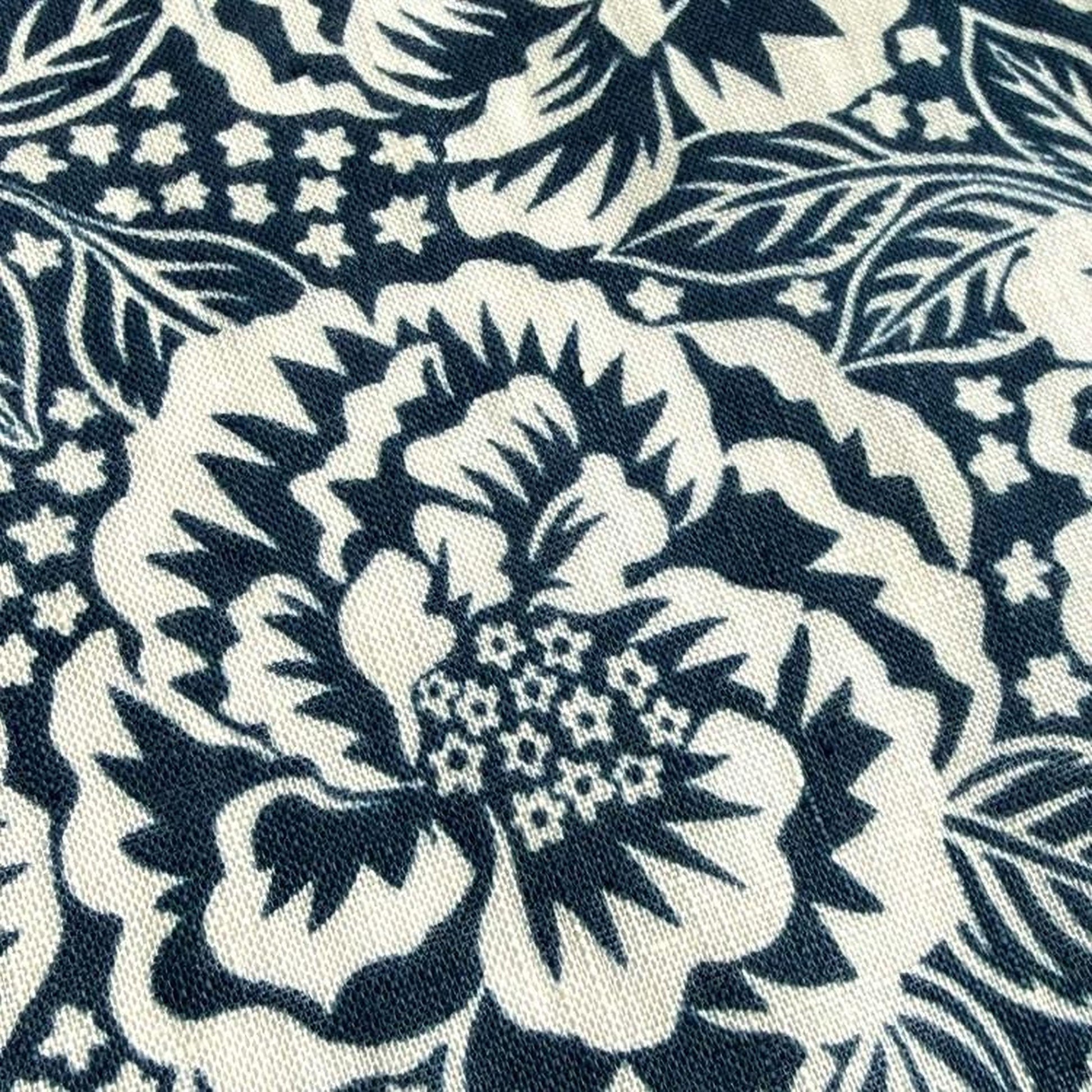 a close-up view of a fabric with a black and white floral pattern. The pattern consists of large, stylized flowers with intricate details and small white flowers scattered throughout the design. The fabric appears to be a dark-colored material, possibly a type of cotton or linen, with a textured surface.