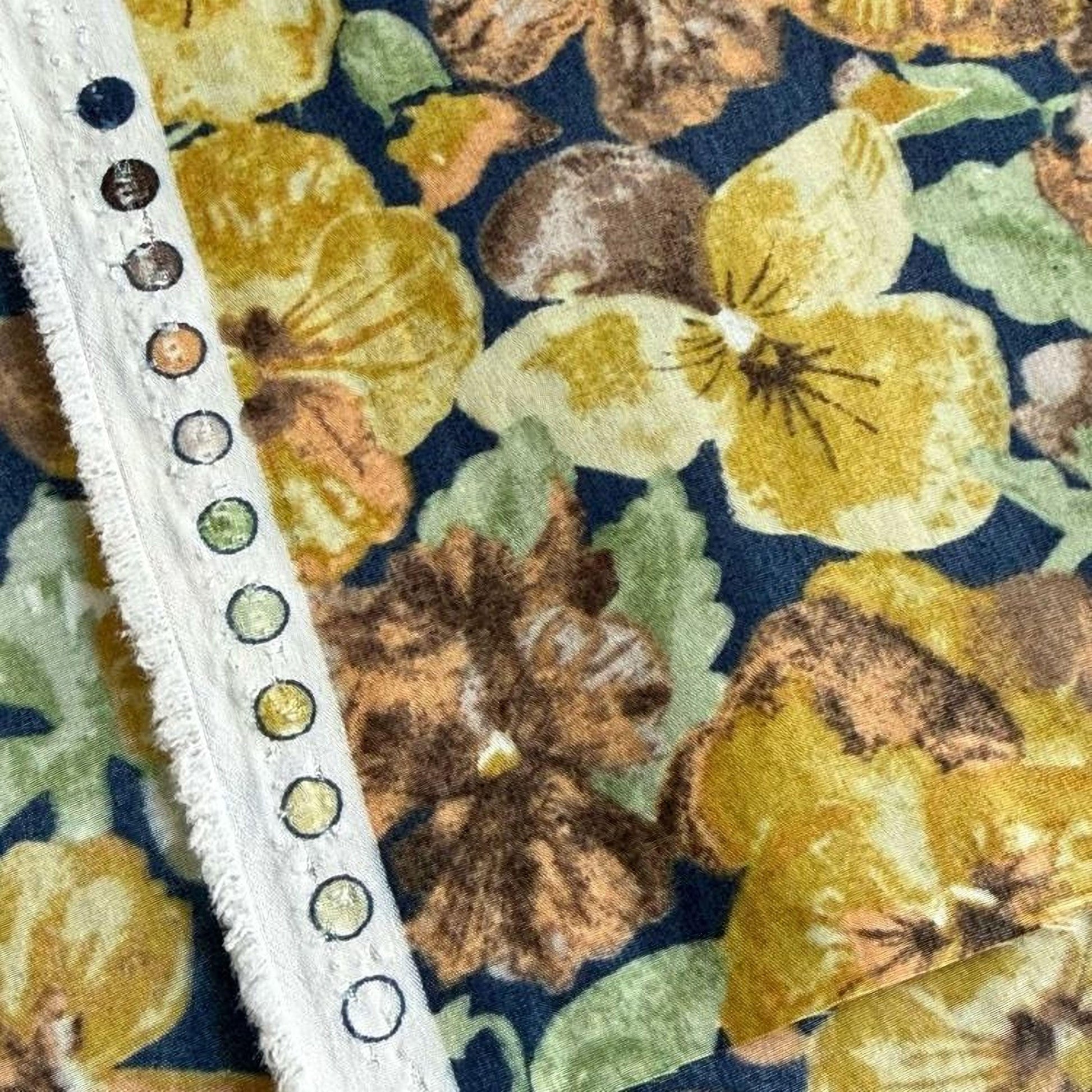a close-up view of a fabric with a floral pattern, including yellow and brown flowers. The fabric appears to be a dark blue or navy blue background with a white fringe or trim running along the edge.