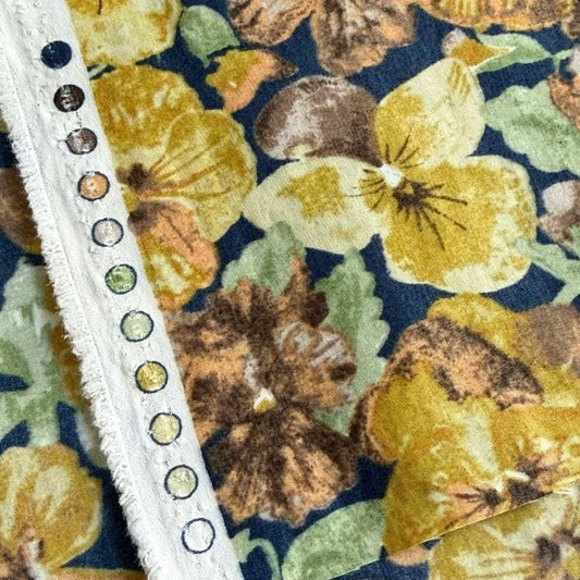 a close-up view of a fabric with a floral pattern, including yellow and brown flowers. The fabric appears to be a dark blue or navy blue background with a white fringe or trim running along the edge.