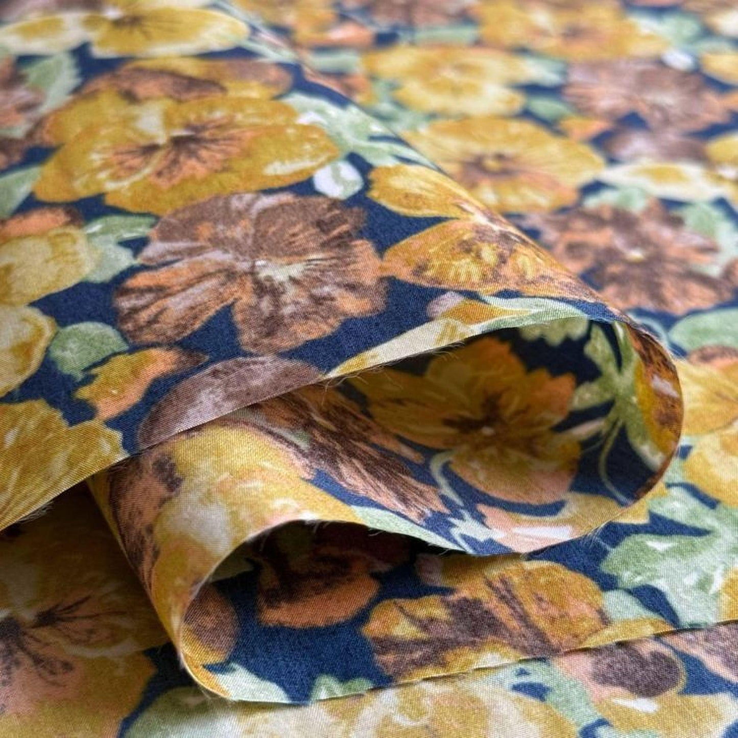 a close-up view of a fabric with a vibrant floral pattern in shades of yellow, orange, and brown. The fabric appears to be folded or rolled, revealing the intricate design of the flowers.