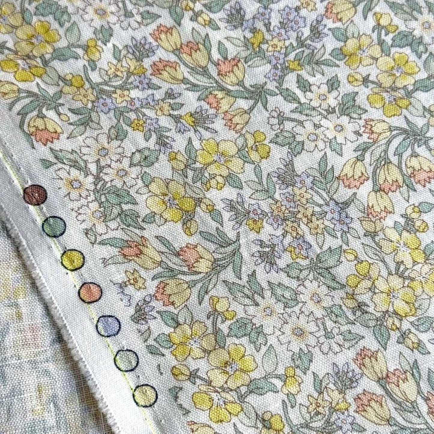 a close-up view of a fabric with a floral pattern, featuring yellow and white flowers and leaves. The fabric appears to be a piece of cloth or textile, with a colorful border or trim visible on the left side.