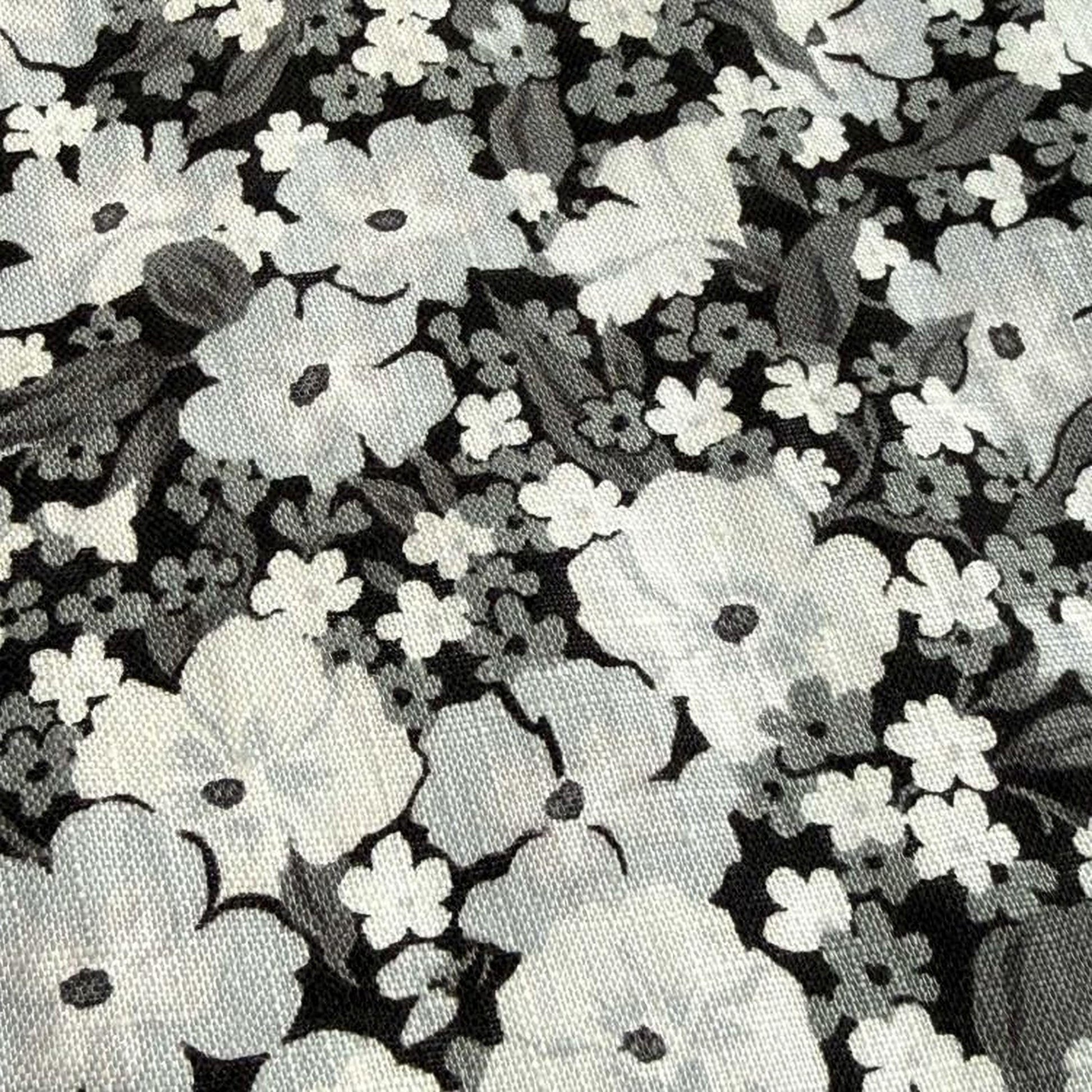 a close-up view of a fabric with a black and white floral pattern. The fabric appears to be a type of textile, possibly a piece of clothing or a decorative item. The pattern consists of numerous white flowers interspersed with black leaves, creating a visually striking contrast against the dark background. The image does not contain any text or additional objects, and the focus is solely on the intricate floral design.