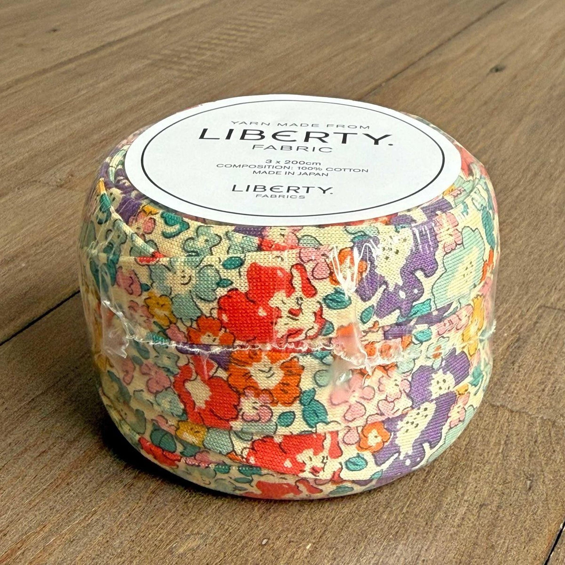 A package of Liberty fabric, featuring a colorful floral pattern, is displayed on a wooden surface.