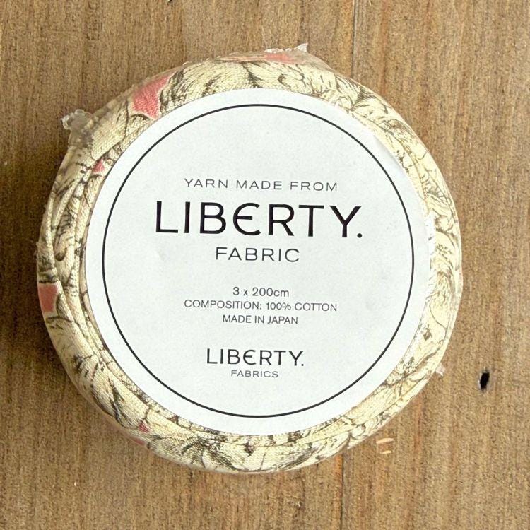 A circular container with a label that reads "Yarn Made from LIBERTY. Fabric" and provides details about the product, including its composition and manufacturing origin.