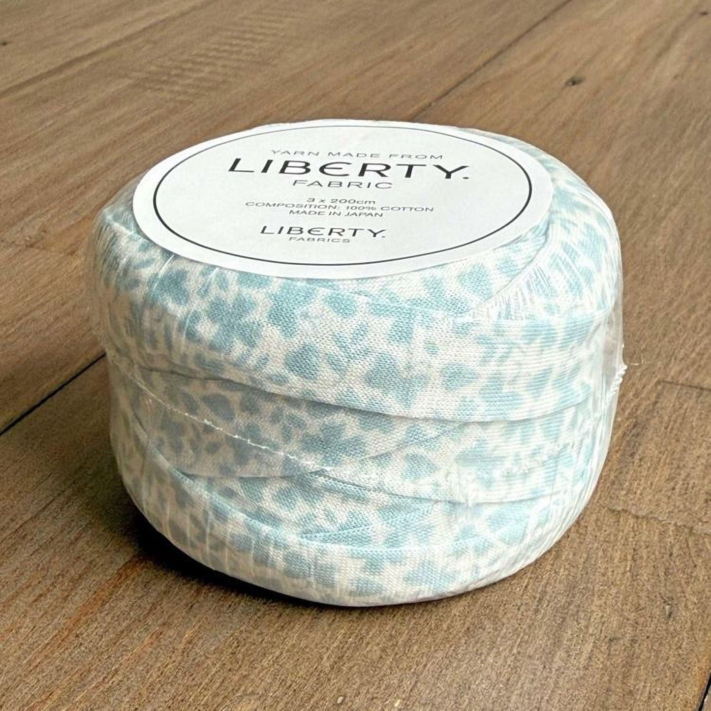 A roll of blue and white fabric with a label that reads "Yarn Made from LIBERTY Fabric" is sitting on a wooden surface.