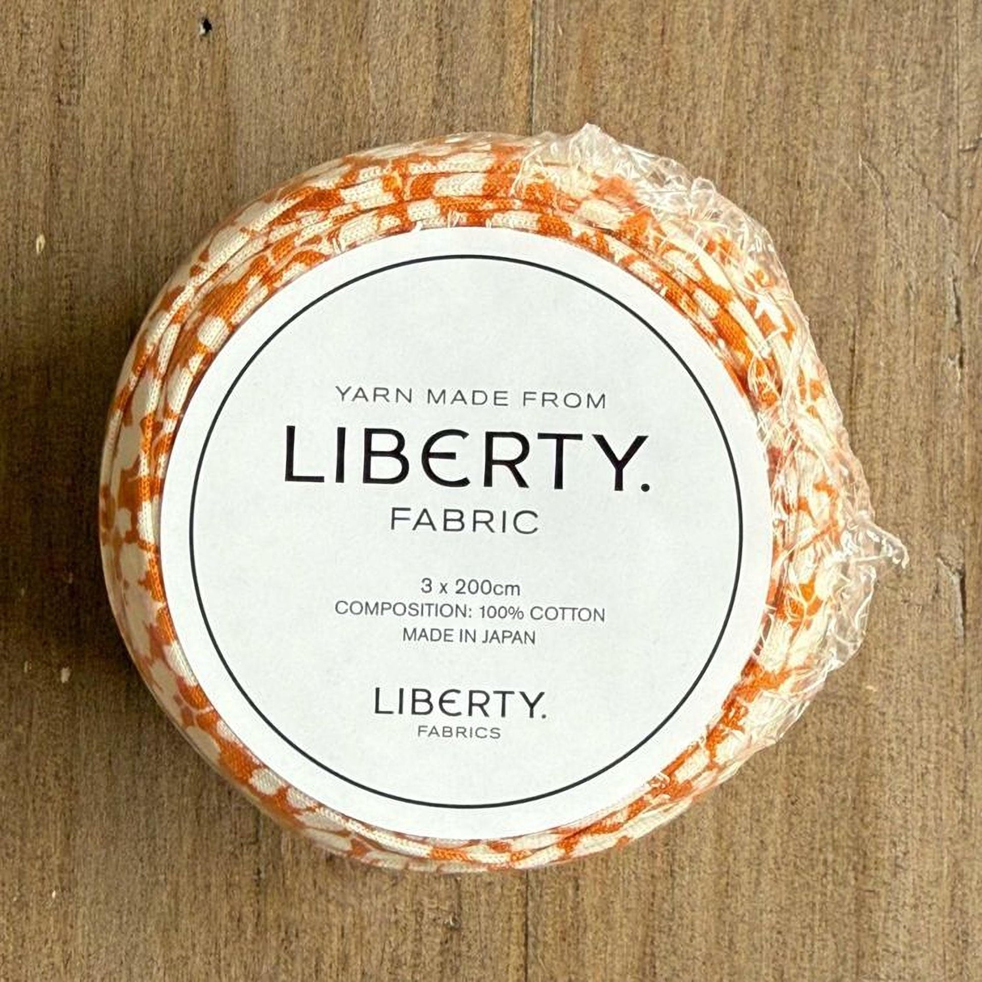 A white label with black text on a wooden surface, displaying the text "YARN MADE FROM LIBERTY FABRIC" and "3 x 200cm COMPOSITION: 100% COTTON MADE IN JAPAN LIBERTY FABRICS".