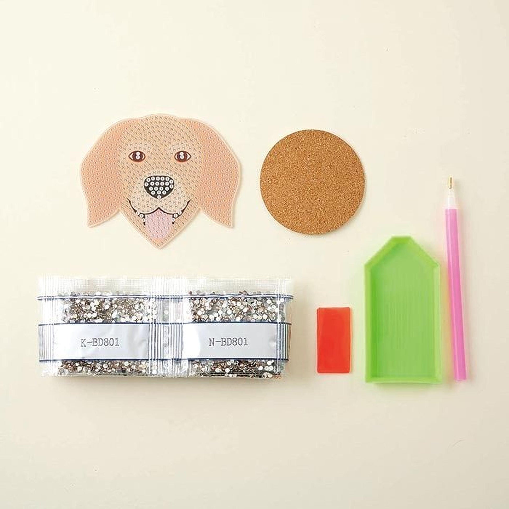 various craft supplies, including a dog-shaped paper, a cork coaster, a pen, a small bag of glitter, and a diamond-studded tool.