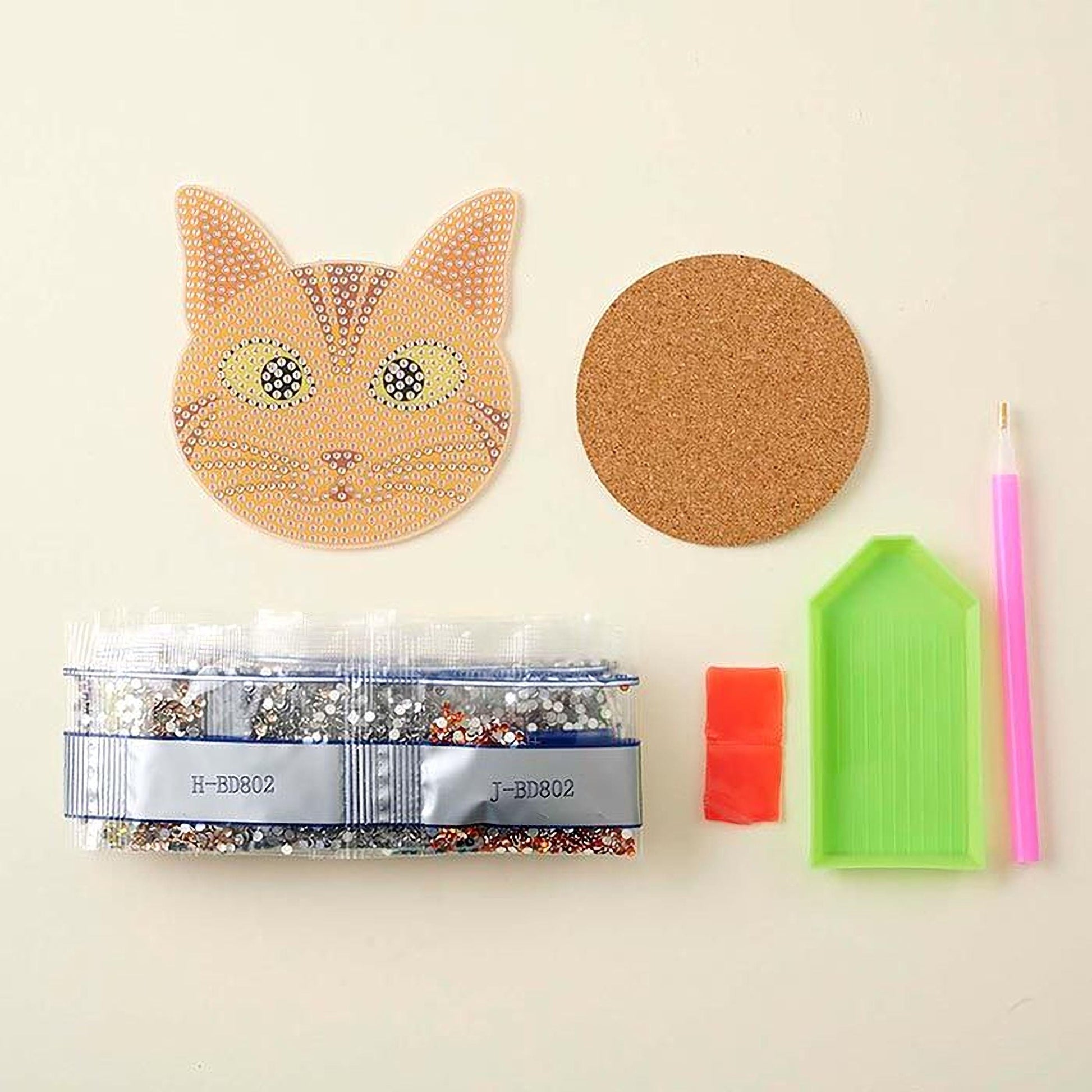 various craft supplies, including a cat-shaped object, a cork coaster, a pen, a clear plastic bag filled with glitter, and a diamond-studded object.
