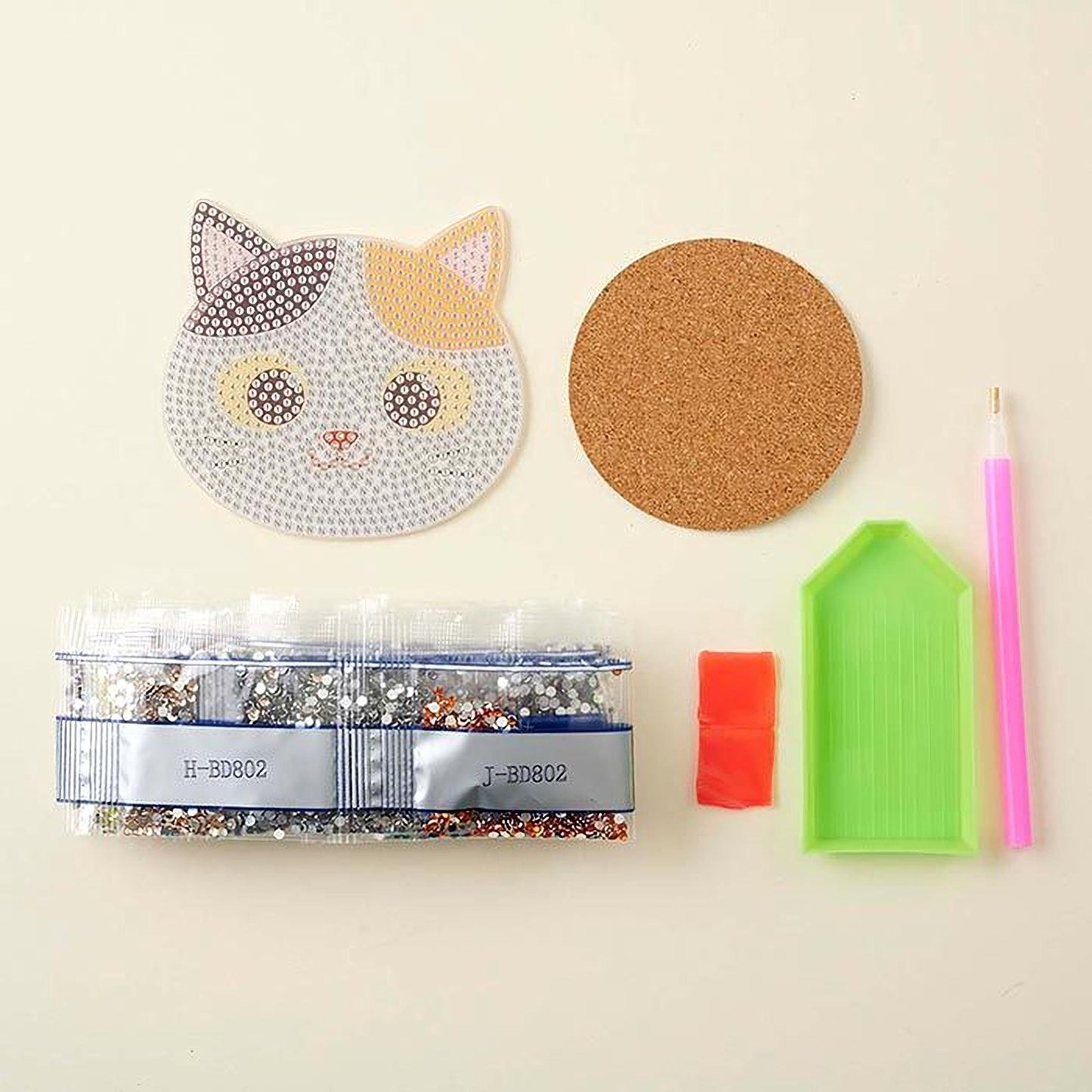 various craft supplies, including a cat-shaped object, a cork coaster, a pen, a small green tray, and a package of rhinestones.