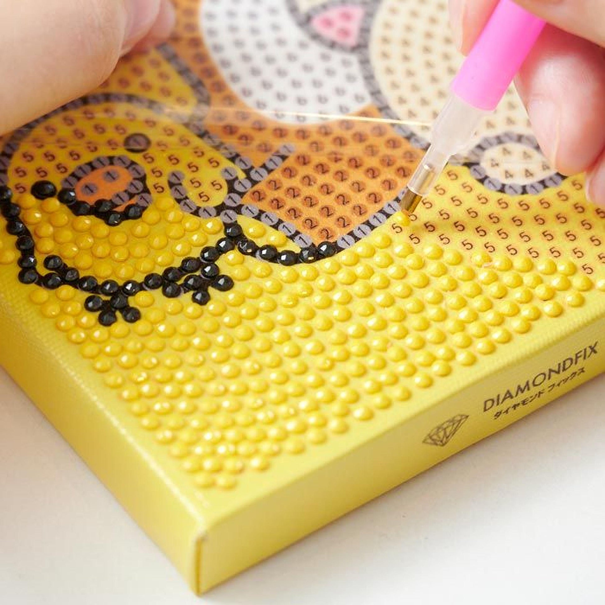 A person's hand is using a pink pen to work on a yellow canvas with a pattern of small dots, likely for a creative project or art activity.