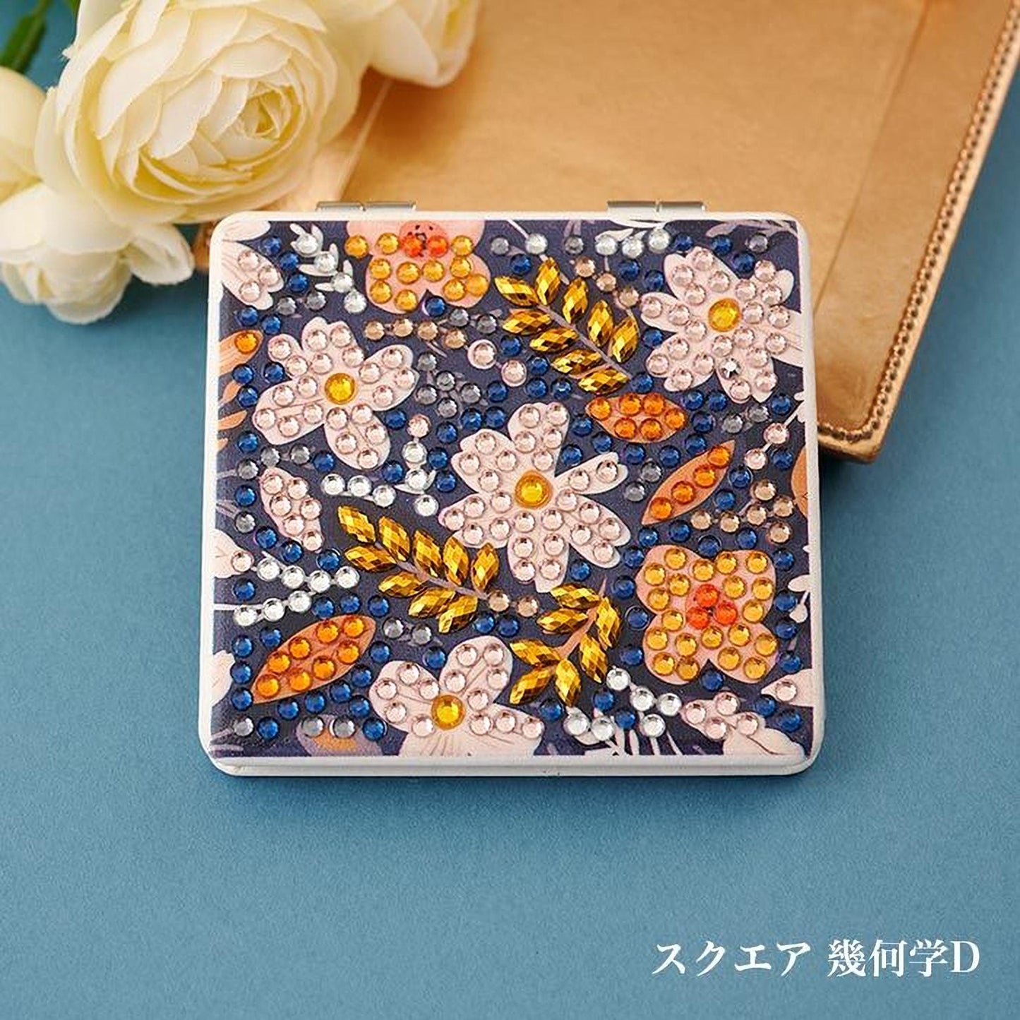 A decorative compact mirror with a floral pattern and rhinestones, placed on a blue surface next to a bouquet of white roses.