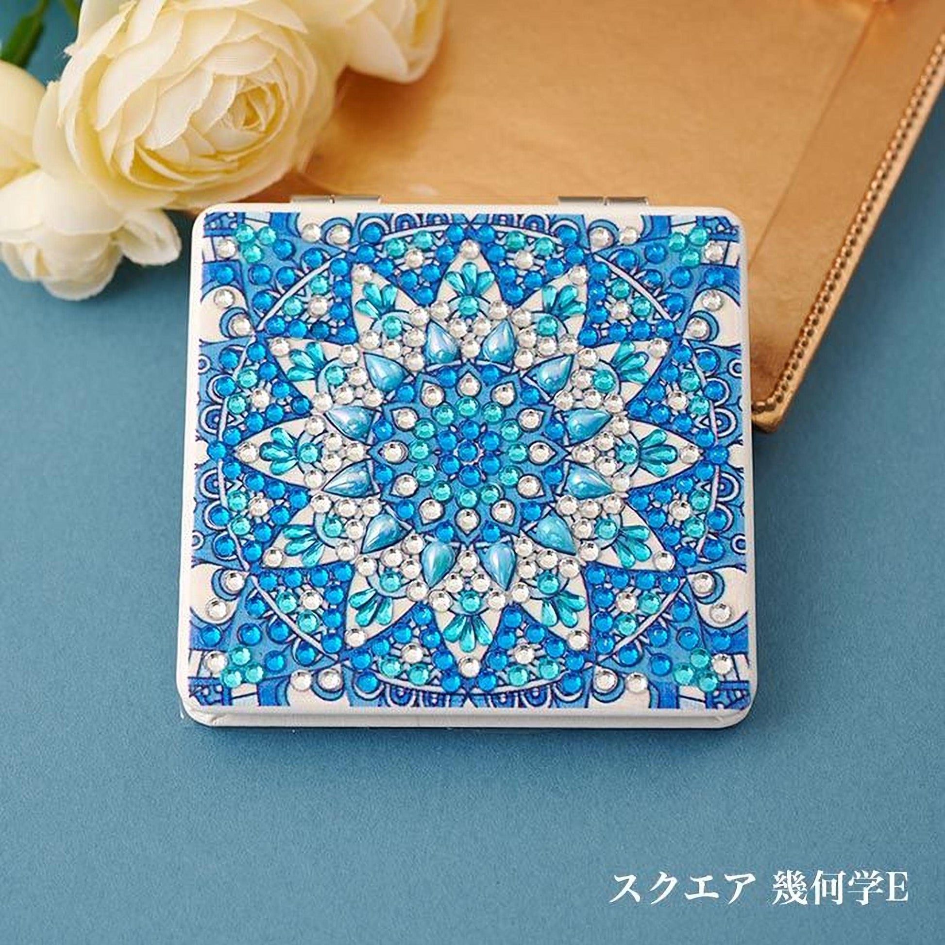 A square-shaped compact mirror with a blue and white floral design, featuring numerous small blue and white gemstones. The mirror is placed on a blue surface, with a bouquet of white roses in the background.