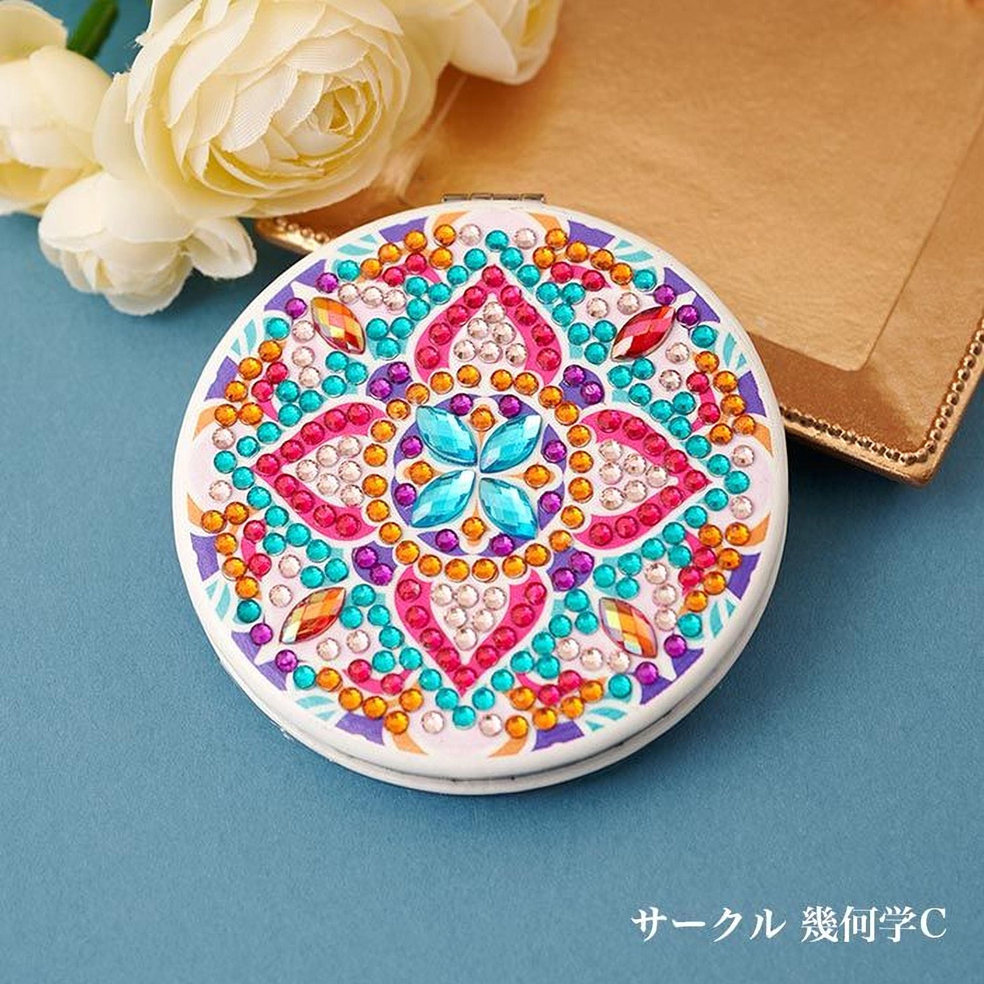 A round mirror with a colorful, intricate design of jewels and flowers, placed on a blue background next to a bouquet of white flowers.