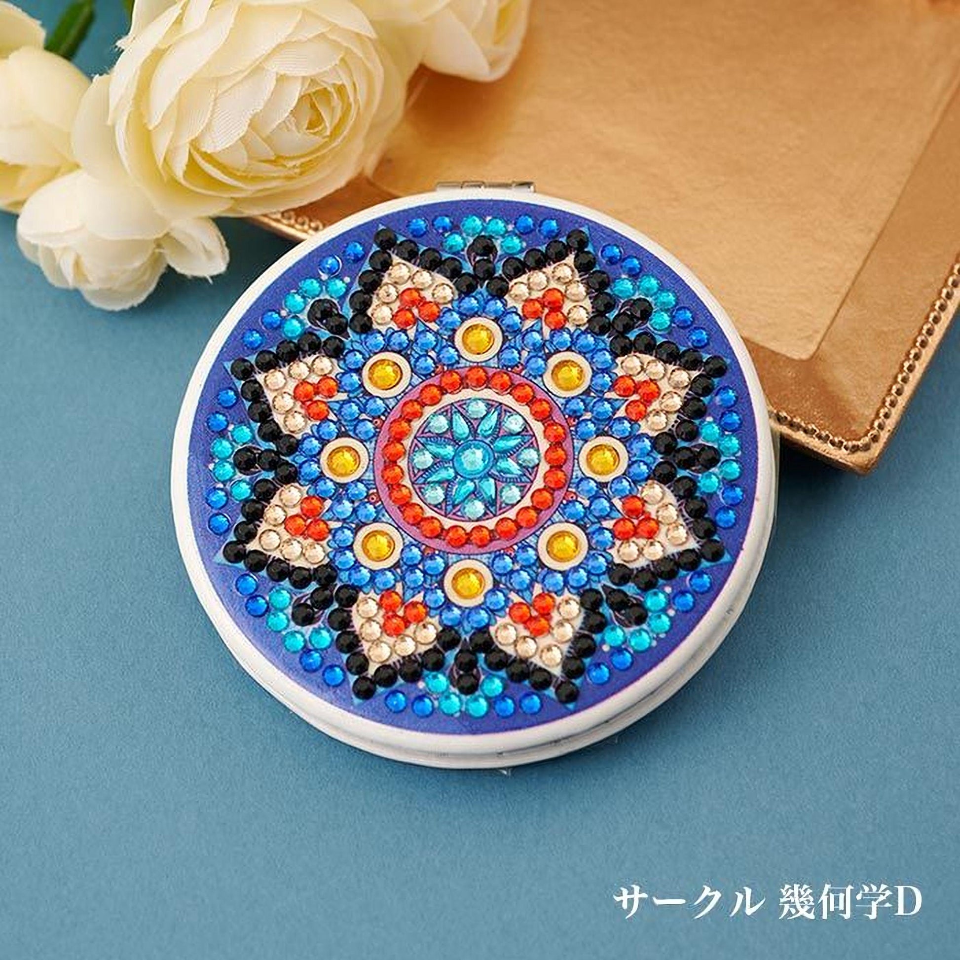 A round mirror with a blue and red patterned design, featuring a central flower-like shape surrounded by smaller geometric shapes and colorful beads. The mirror is placed on a blue surface, with white flowers visible in the background.