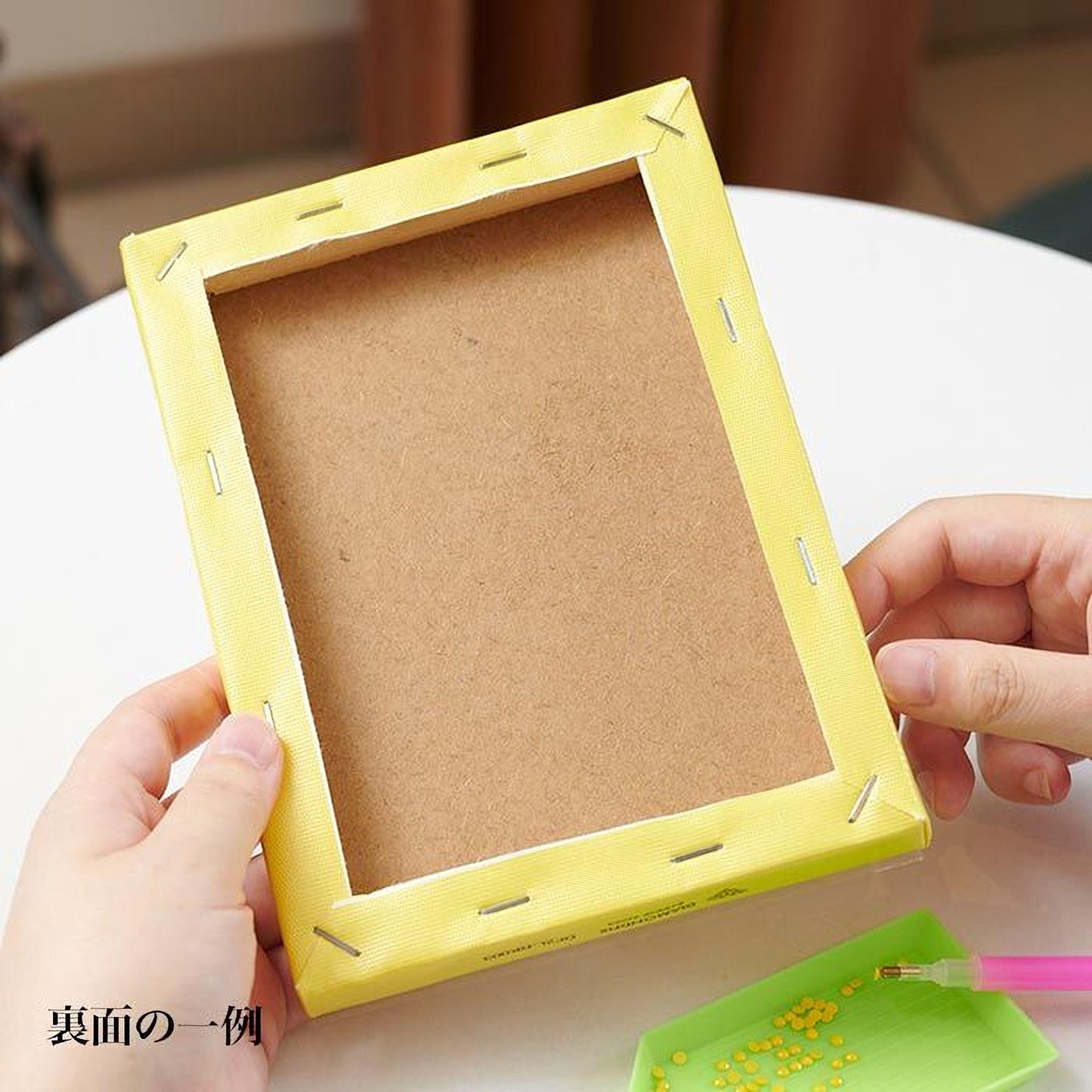 A person's hands are holding a yellow frame with a blank canvas inside, suggesting they are in the process of creating a piece of art or craft.