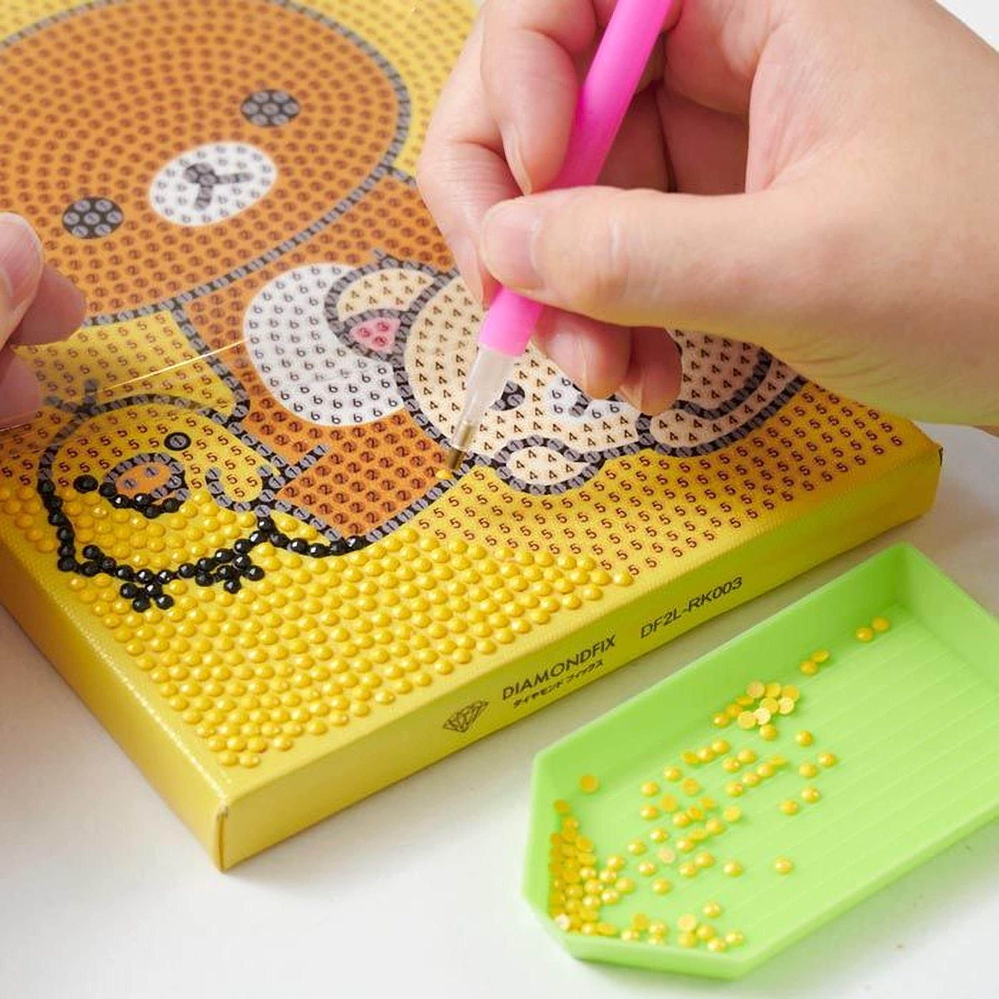 A person's hands are carefully placing small yellow beads onto a yellow canvas, which is decorated with a pattern of black dots and a brown bear.
