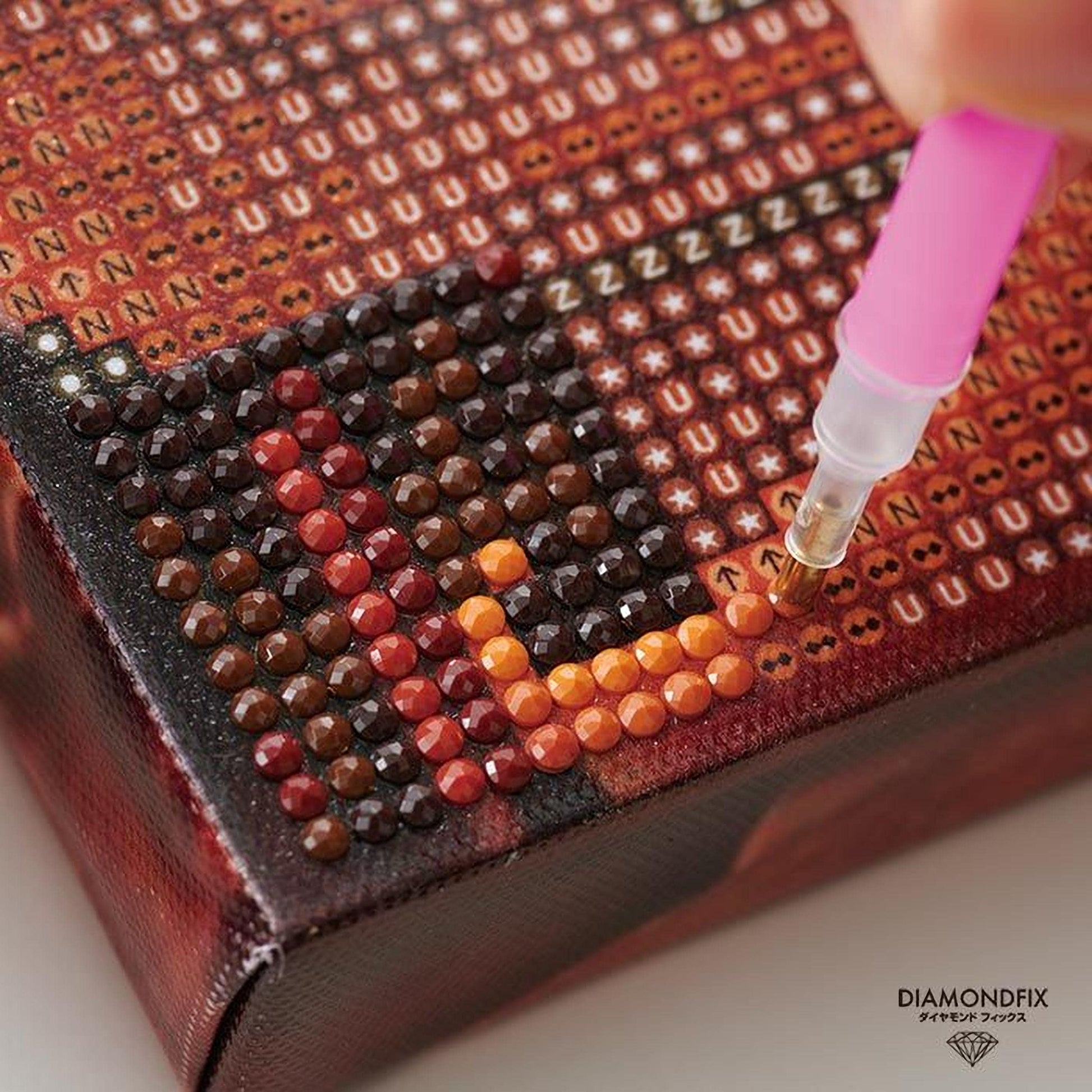 A close-up image of a person using a pink pen to carefully apply rhinestones to a canvas or surface, creating a detailed and intricate design.