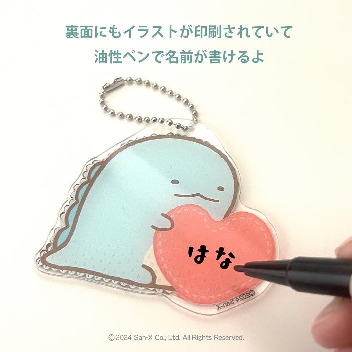 a keychain with a small blue fish and a heart-shaped charm attached to it. A black pen is being used to write on the charm, which has the Japanese characters "おとなしい" (heart).