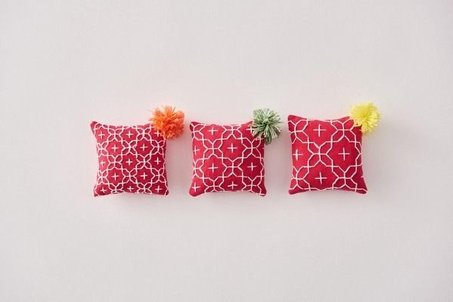 Three red and white patterned pillows with pom-poms are arranged in a row on a white background.