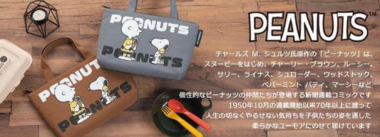 two Peanuts-themed bags, one brown and one gray, placed on a wooden surface alongside various Peanuts-related items such as a plate, a fork, and a spoon.