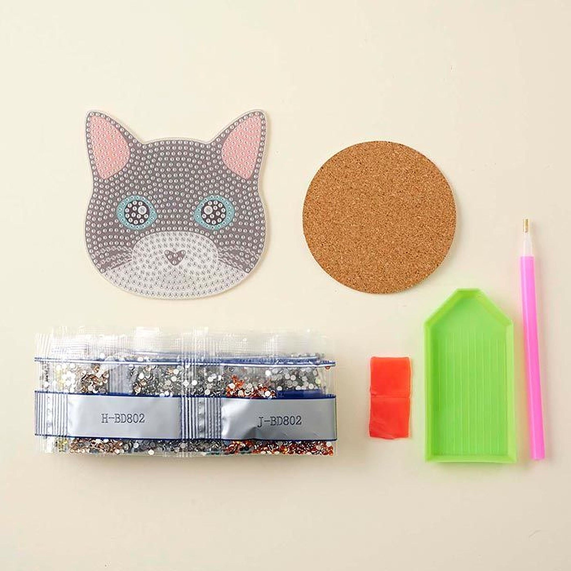 various craft supplies, including a cat-shaped paper, a cork coaster, a pen, a small bag of glitter, and a diamond tool.
