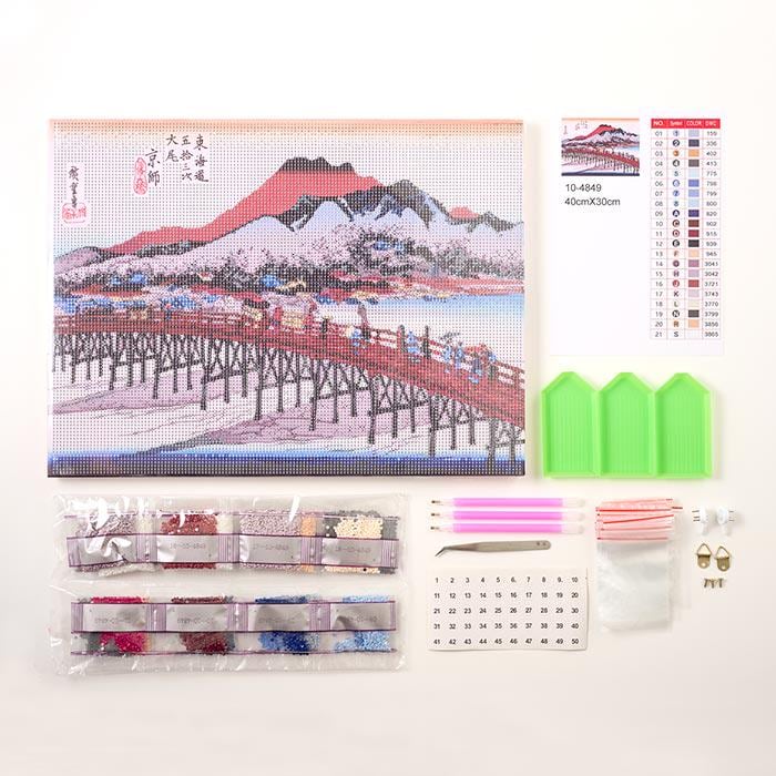 various art supplies and materials, including a large embroidery kit depicting a scenic landscape, a grid for cross-stitching, and a set of tools and beads for creating a cross-stitch design.