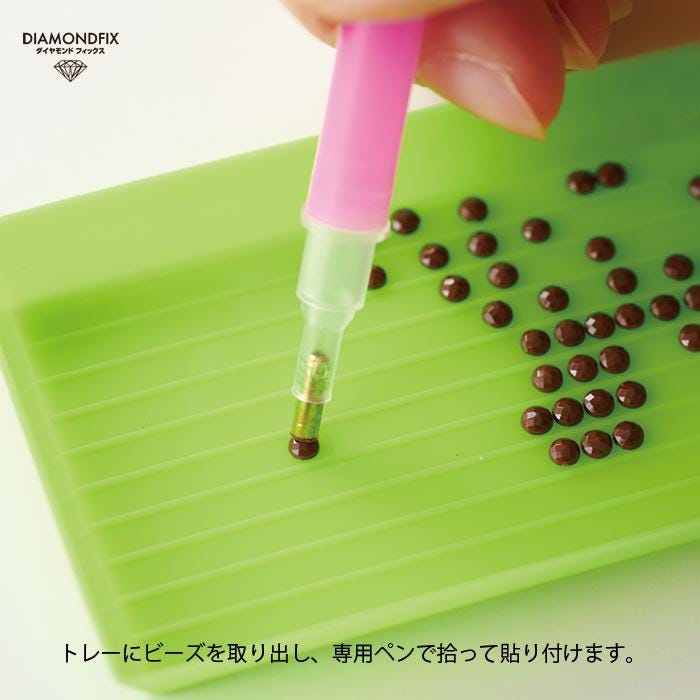A hand is using a pink tool to work on a green surface with numerous small brown objects, likely beads or stones.