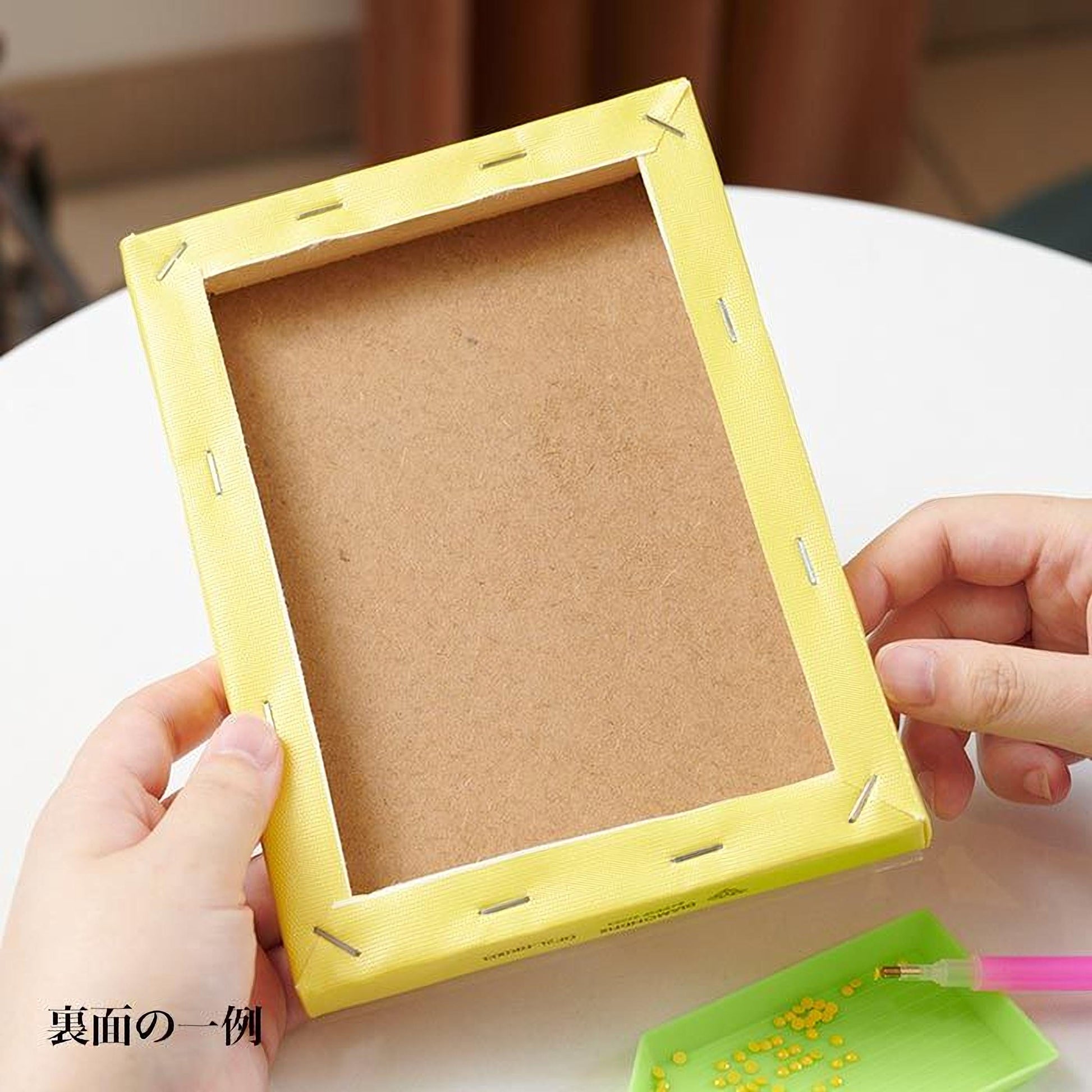 A person's hands are holding a yellow frame with a blank canvas inside, suggesting they are in the process of creating a piece of art or craft.