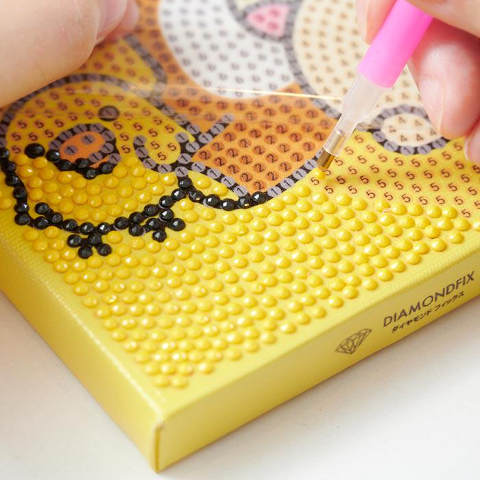 A person's hand is using a pink pen to work on a yellow canvas with a pattern of small dots, likely for a creative project or art activity.