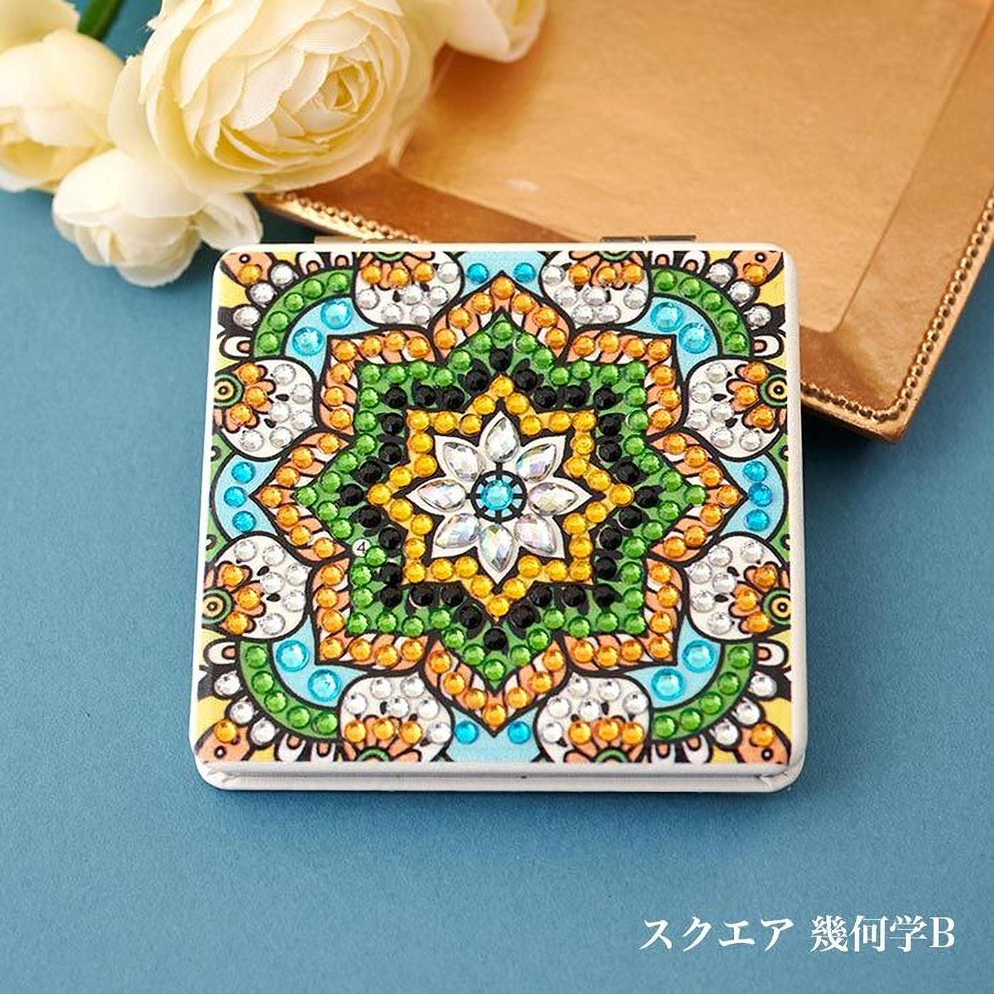 A decorative square mirror with a vibrant, colorful design featuring a central flower-like motif surrounded by intricate patterns and sparkling gemstones. The mirror is placed on a blue surface, with a bouquet of white roses visible in the background.
