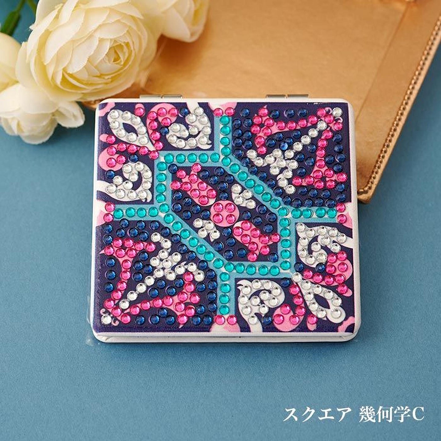A decorative square mirror with a colorful, patterned design made of rhinestones or crystals, placed on a blue surface next to a bouquet of white roses.