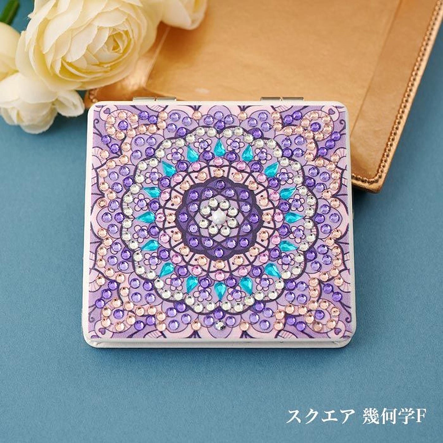 A decorative square-shaped object with a vibrant purple and blue floral pattern, adorned with sparkling jewels, is placed on a blue surface next to a white rose.
