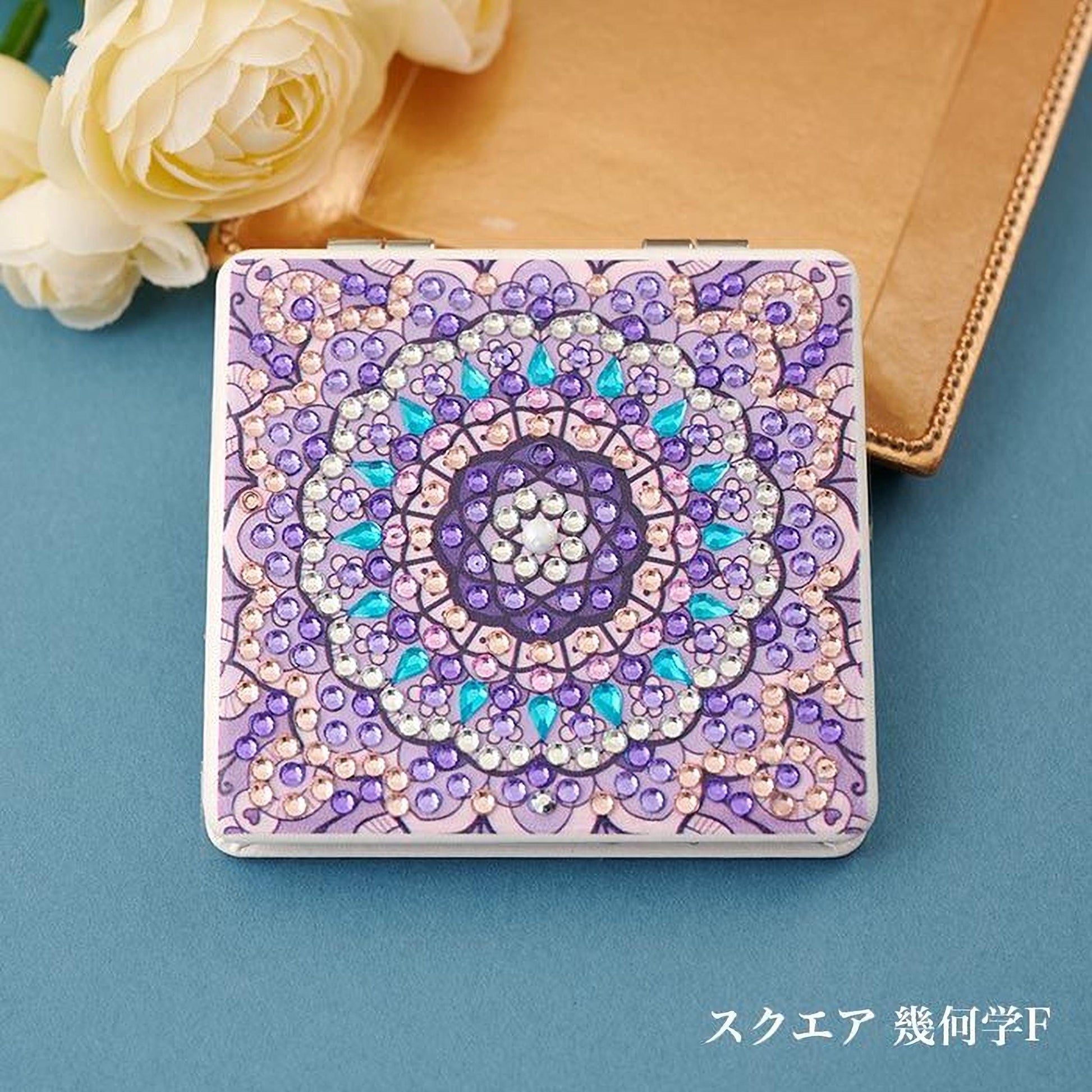 A decorative square-shaped object with a vibrant purple and blue floral pattern, adorned with sparkling jewels, is placed on a blue surface next to a white rose.