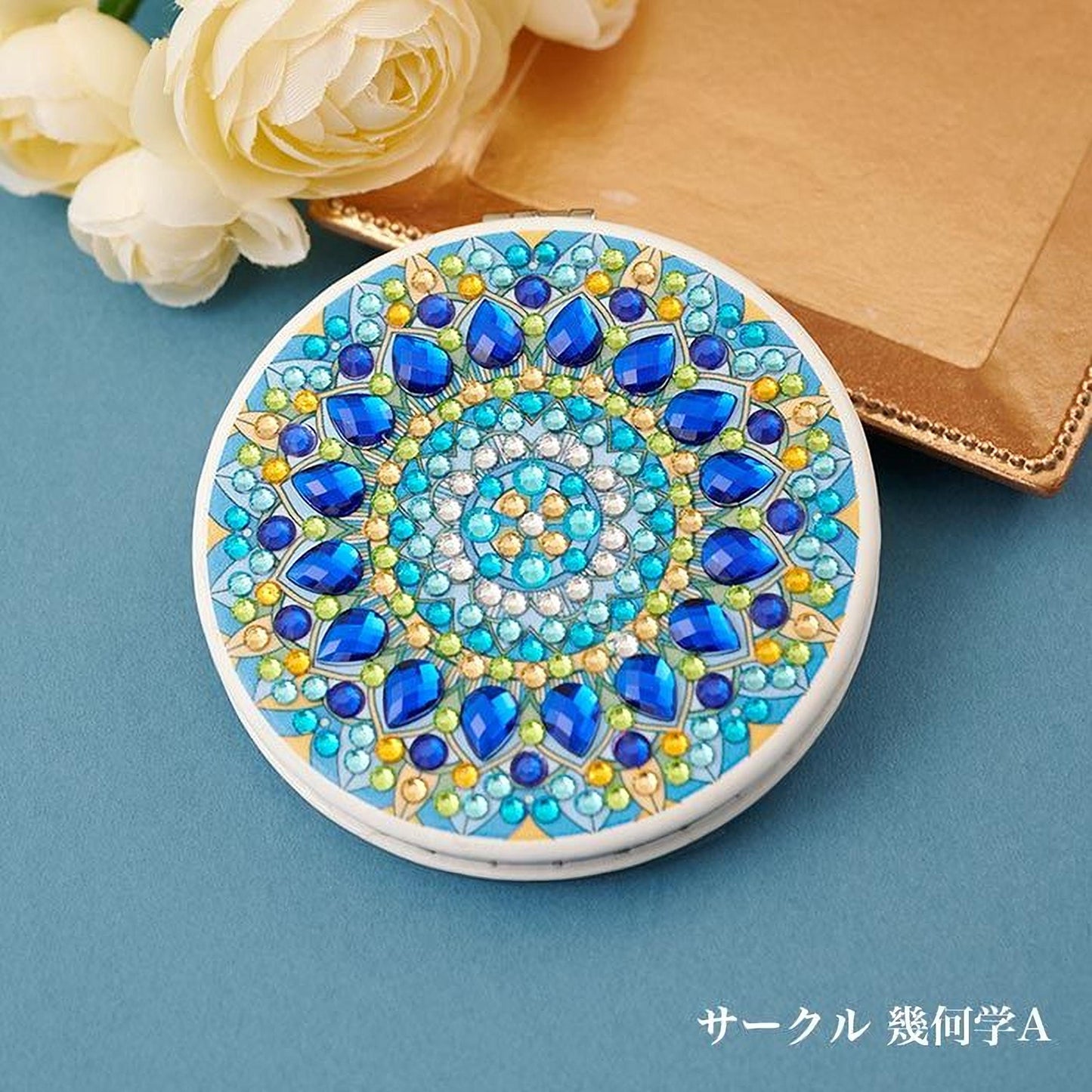 A decorative round mirror with a blue and gold floral design, surrounded by small blue and yellow gemstones, placed on a blue surface next to a bouquet of white flowers.