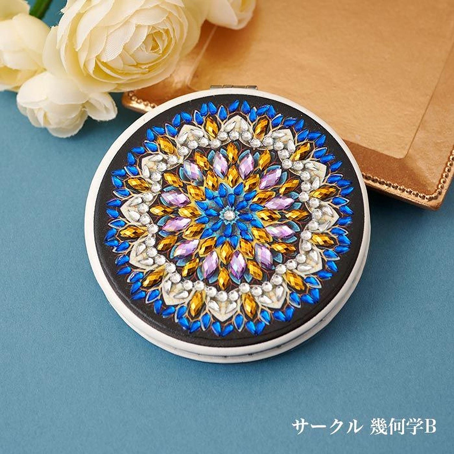 A round mirror with a black frame and a colorful, intricate design of blue, gold, and purple jewels. The mirror is placed on a blue surface, with a bouquet of white flowers in the background.
